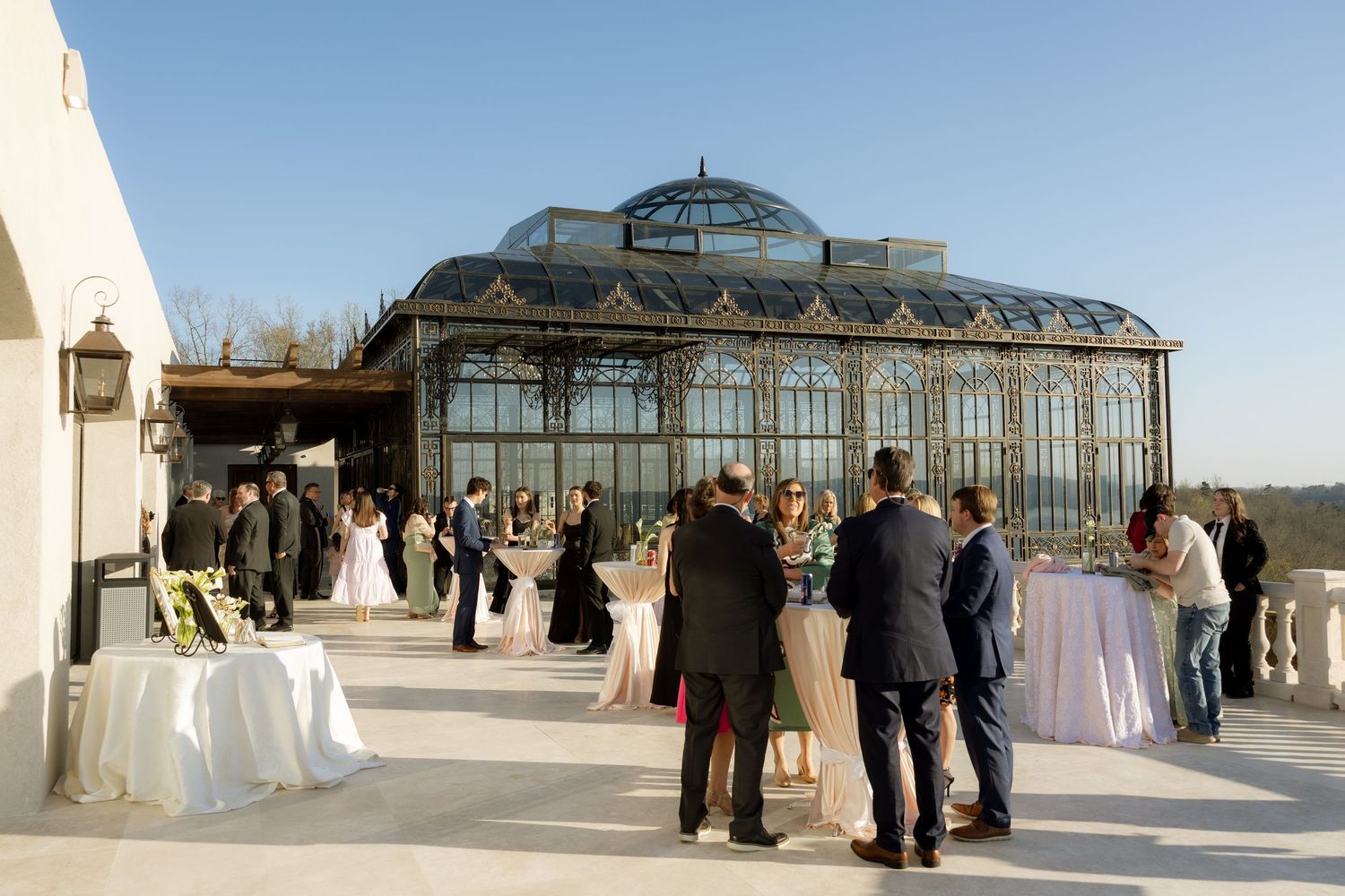 Elegant outdoor wedding reception on a terrace with guests mingling near a grand glass greenhouse venue.