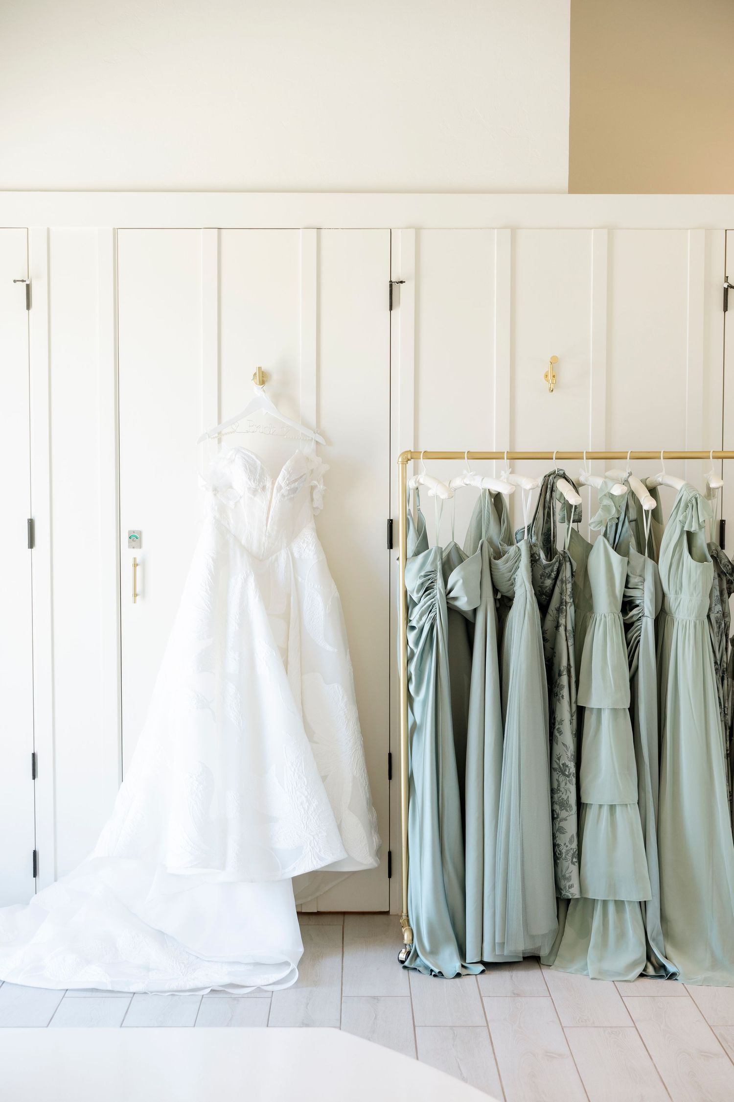 Wedding dresses and sage green bridesmaid gowns hang in a row against a white wall.