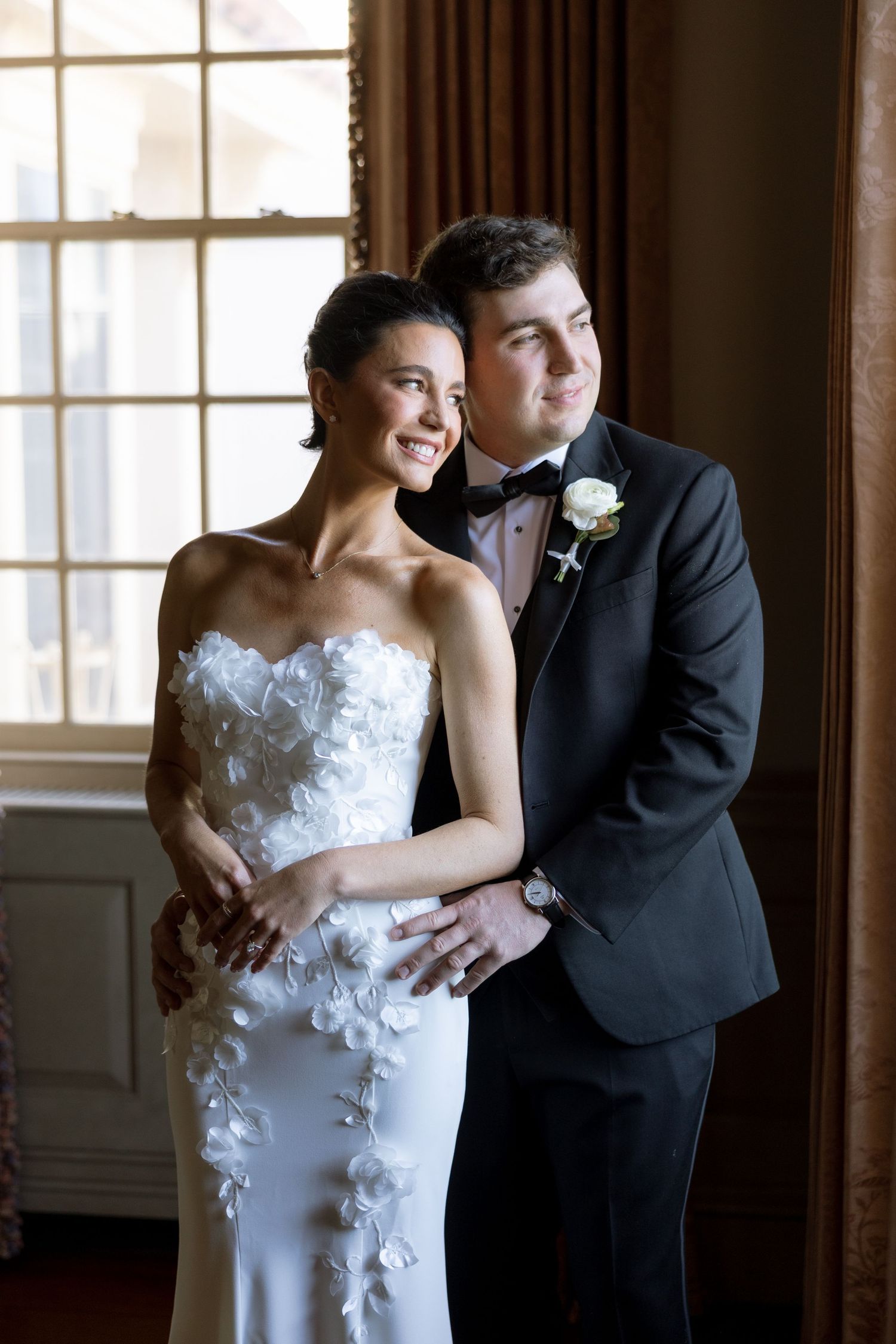 Bride in floral applique strapless gown smiling as groom in black tuxedo embraces her by window.