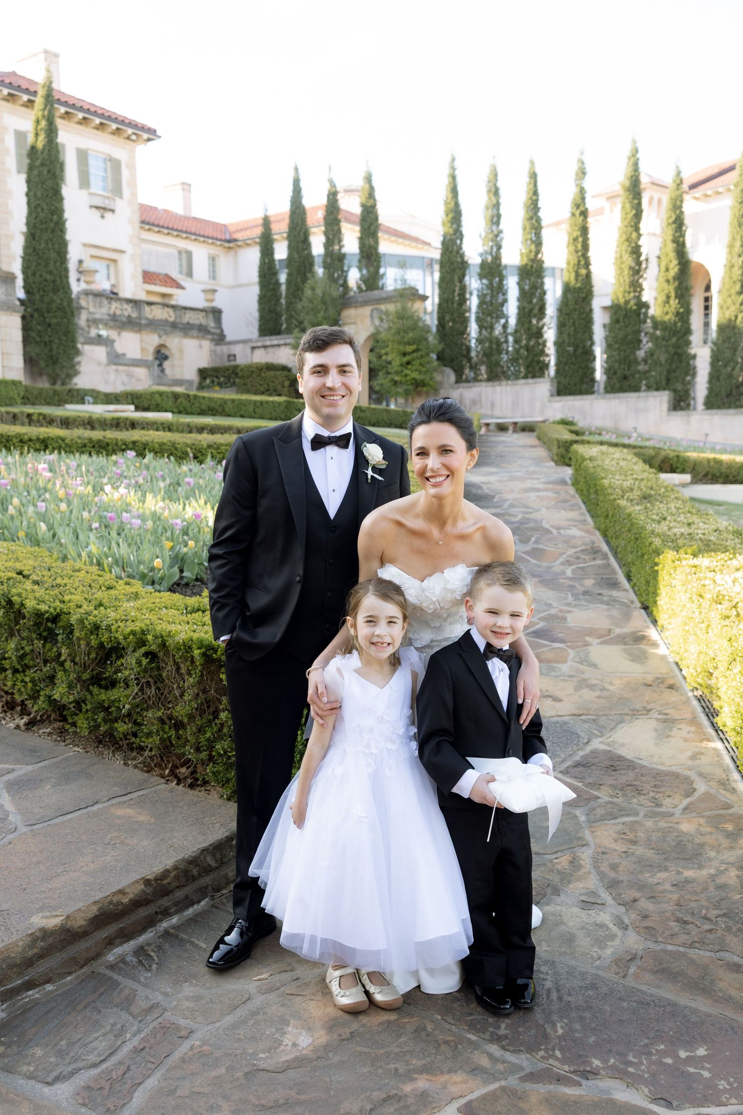 Bride and groom pose with flower girl and ring bearer in tuxedo at elegant garden wedding venue.