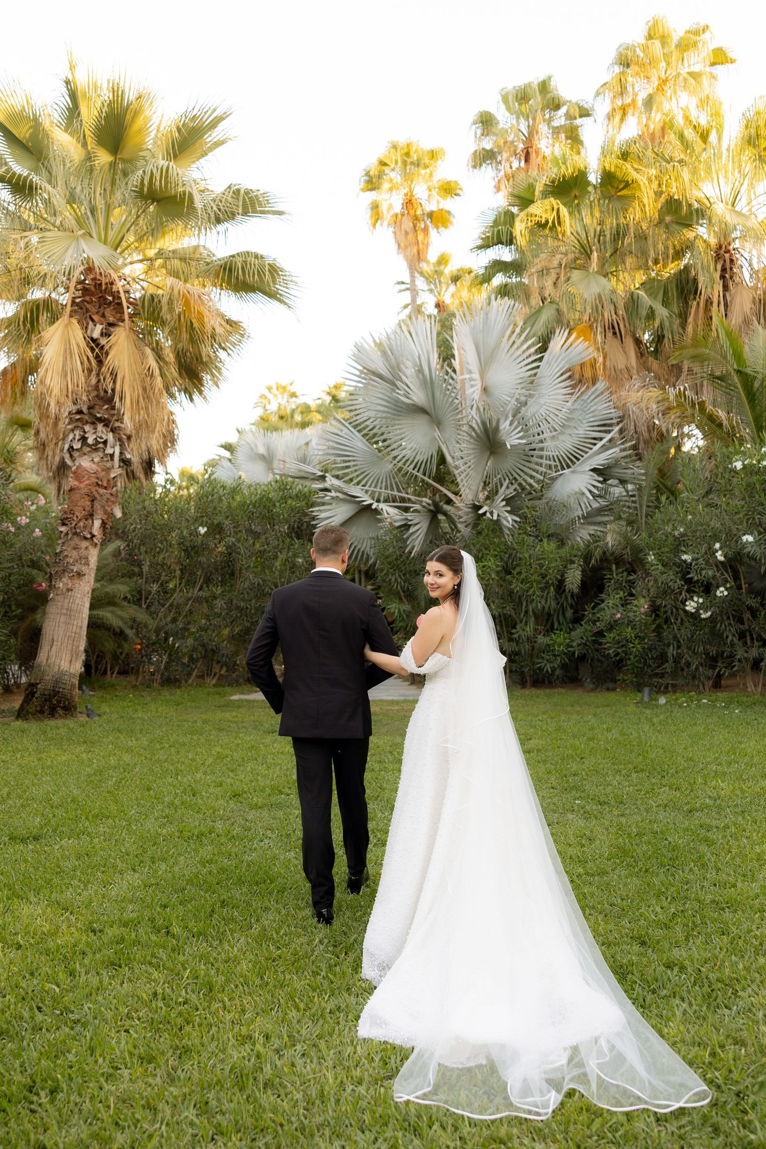 Bride and groom walk hand-in-hand through a lush green garden surrounded by tall tropical palm trees at sunset.