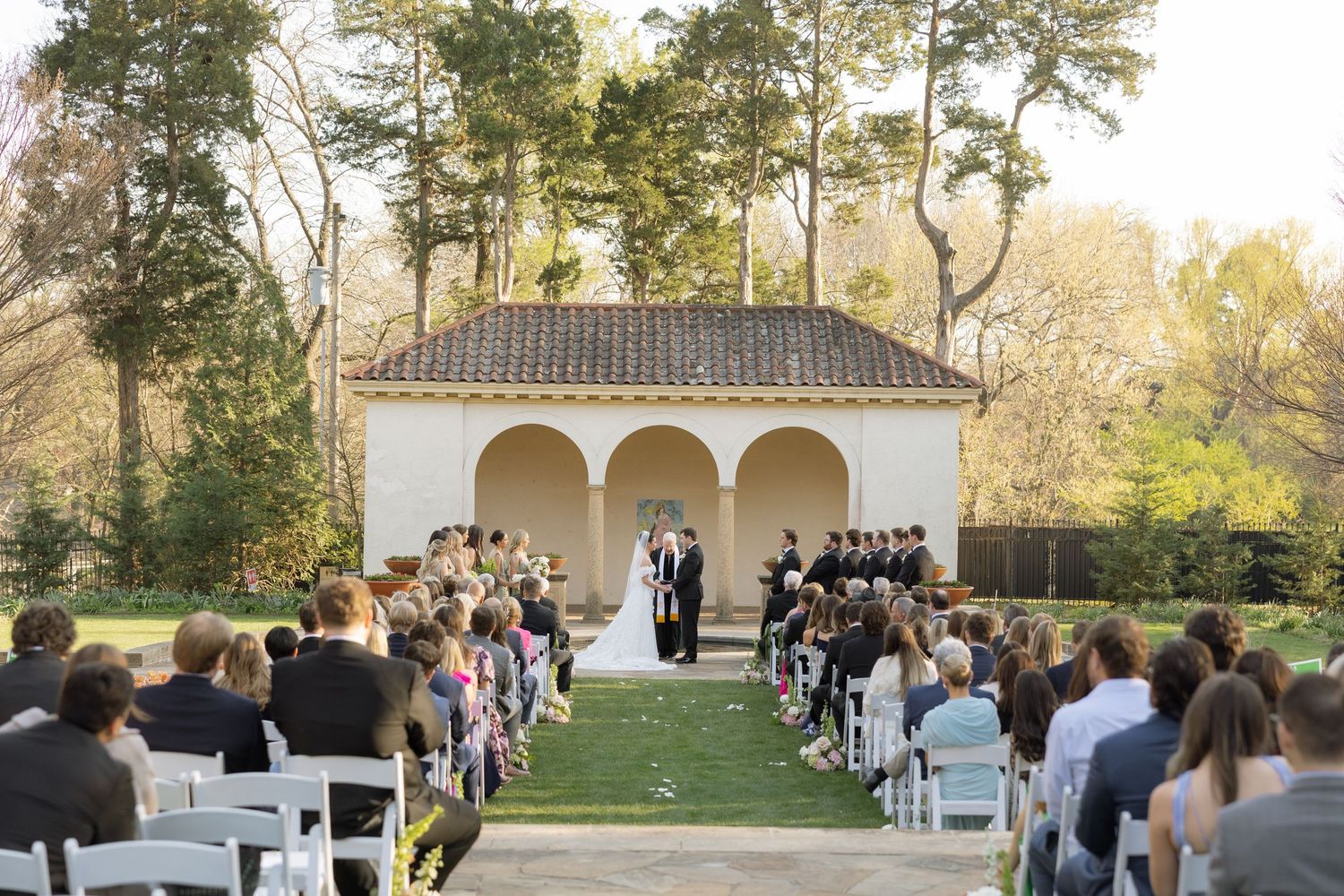 Wedding ceremony on lush green lawn with white chairs and guests seated before a classic arched pavilion.