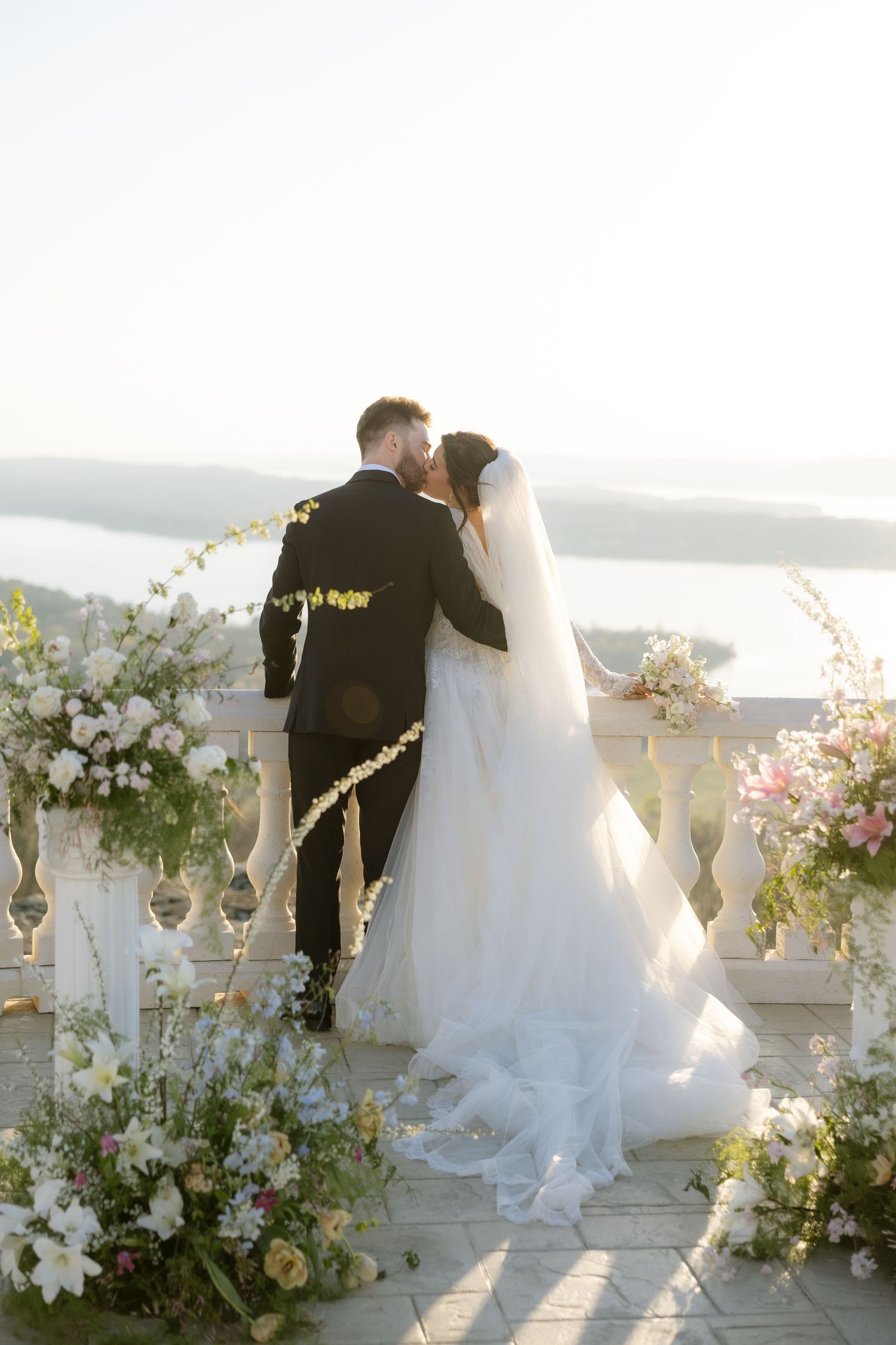 Bride and groom kissing on a floral-adorned balcony with a scenic waterfront view on their wedding day.