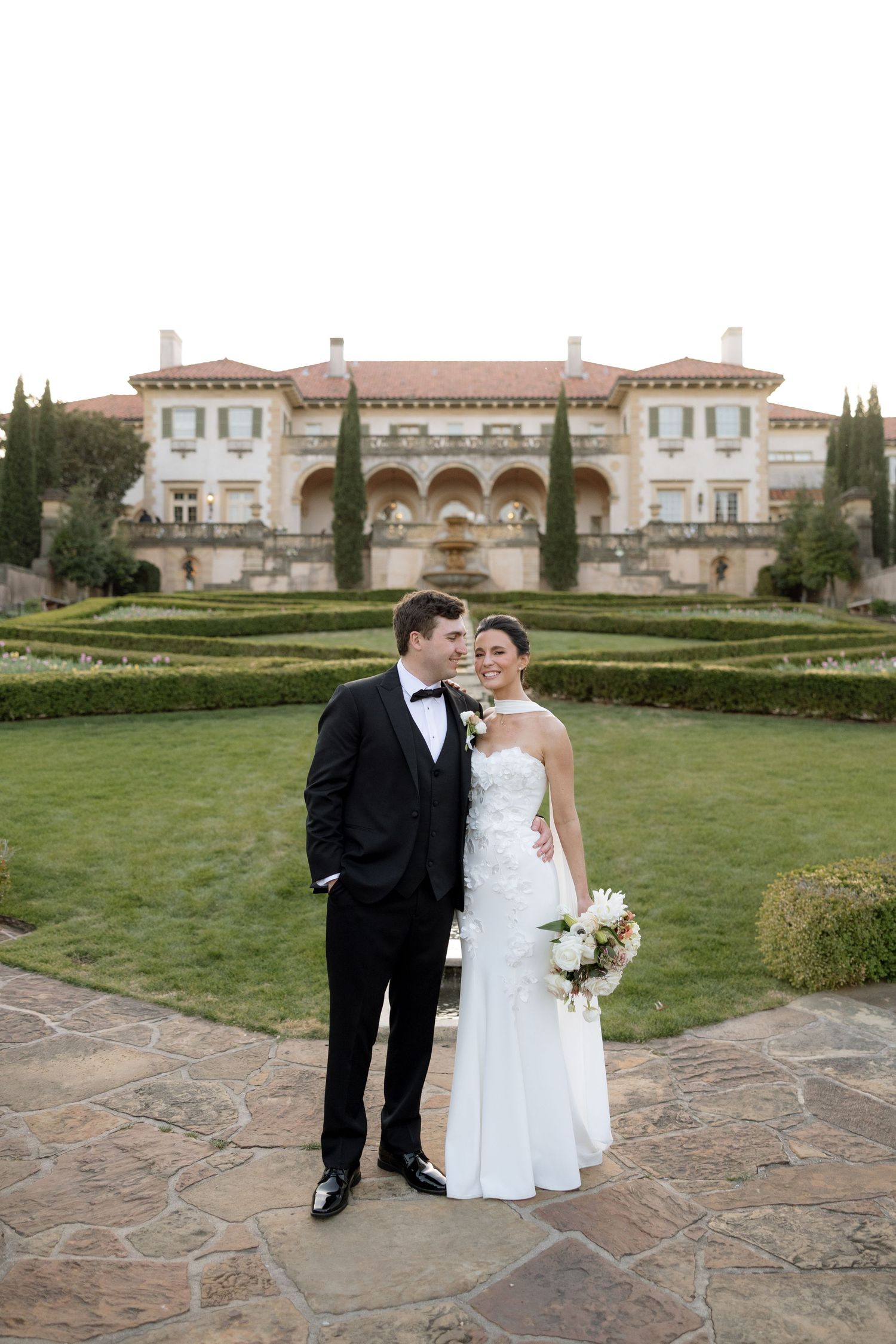 Bride and groom portrait in front of grand Italian-style mansion with manicured gardens.