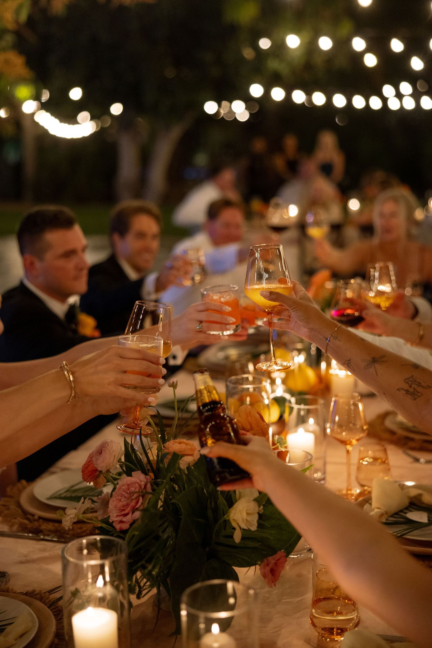 Wedding guests clink glasses of champagne and wine at candlelit outdoor reception under string lights.