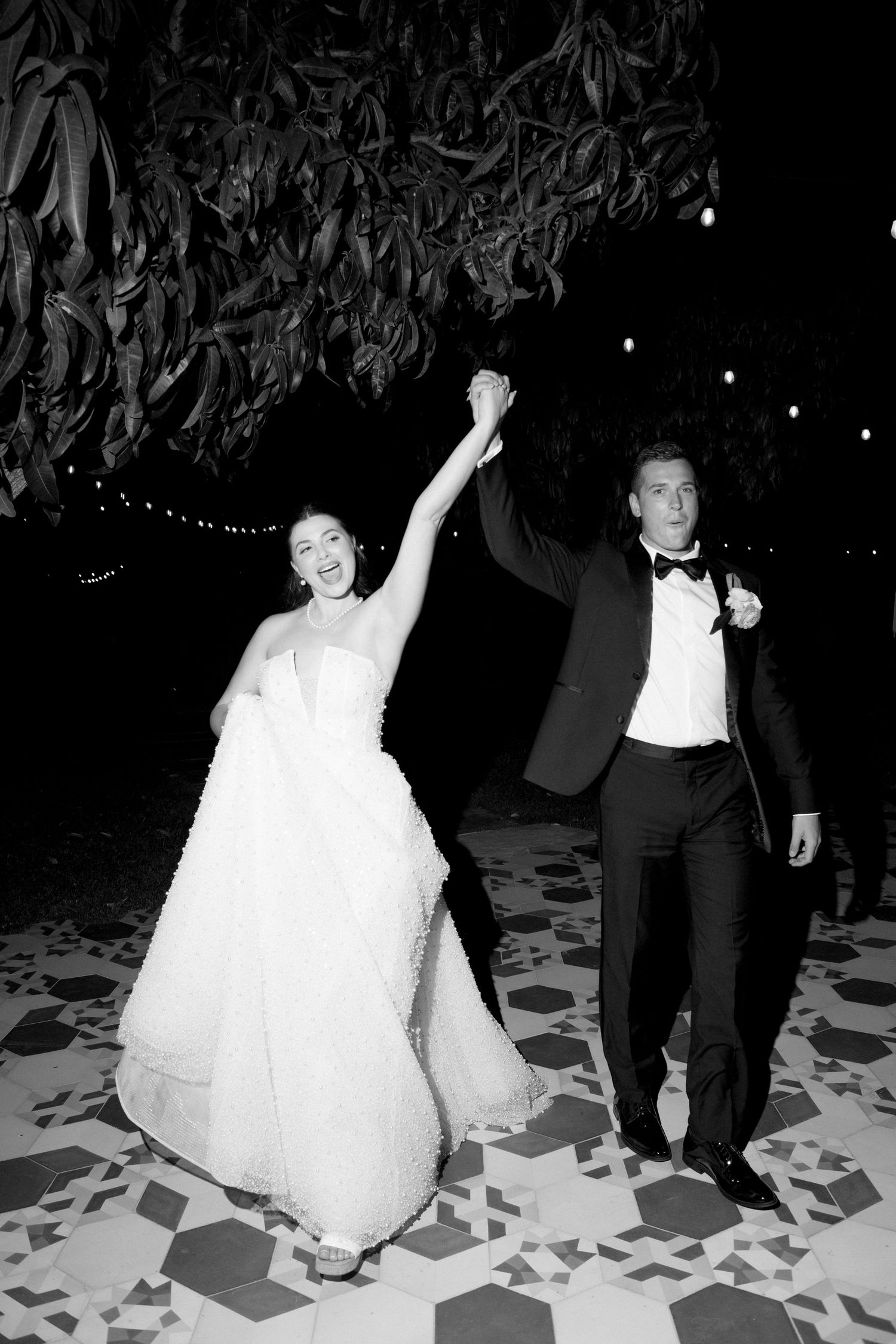 Bride and groom raise hands triumphantly while dancing on patterned floor at night wedding reception.