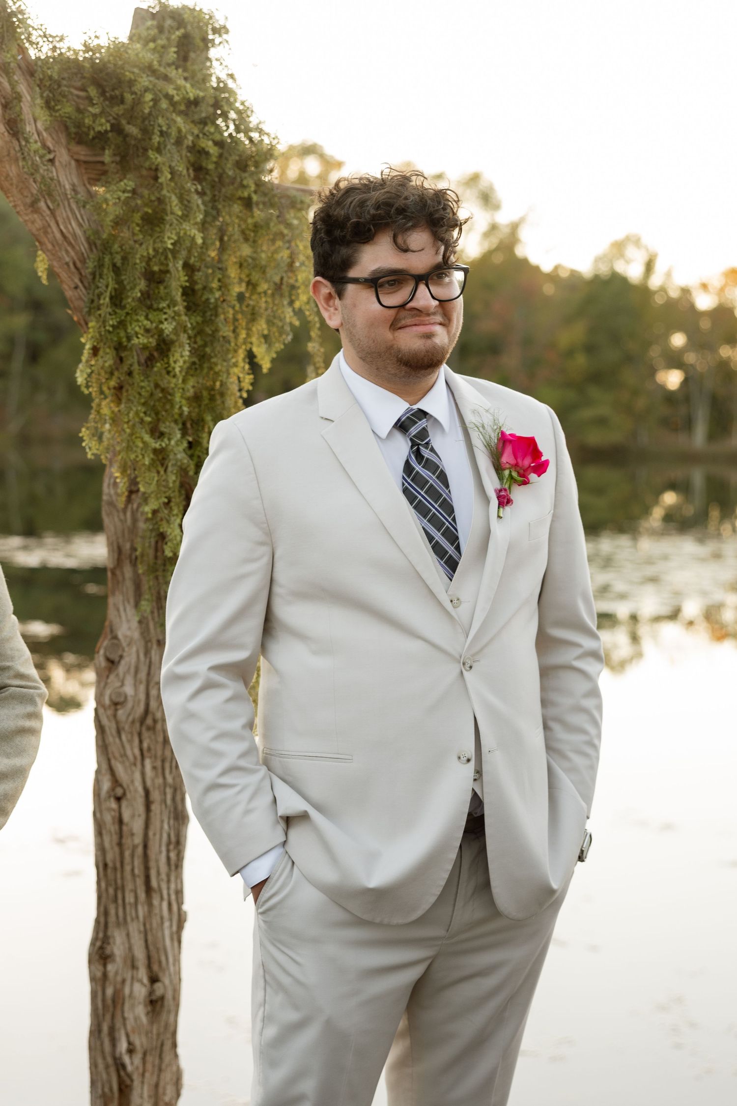 A groom in a light gray suit and glasses stands by a lake at sunset with a pink boutonniere.