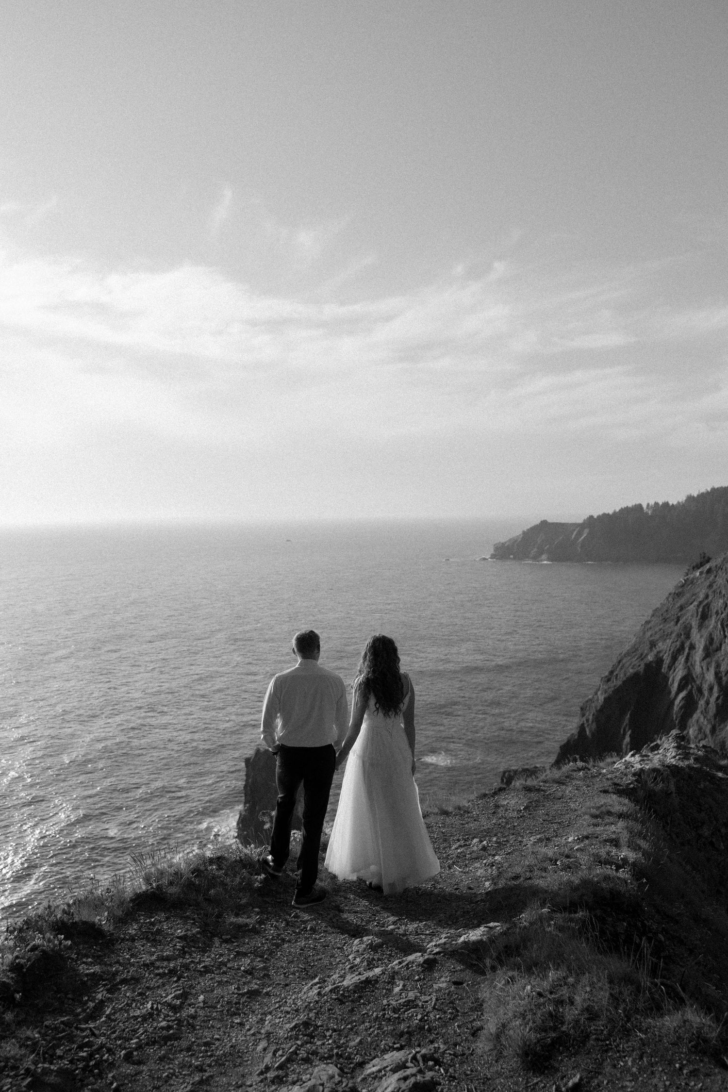 A black and white photo of a couple gazing at the ocean from a dramatic cliff edge.