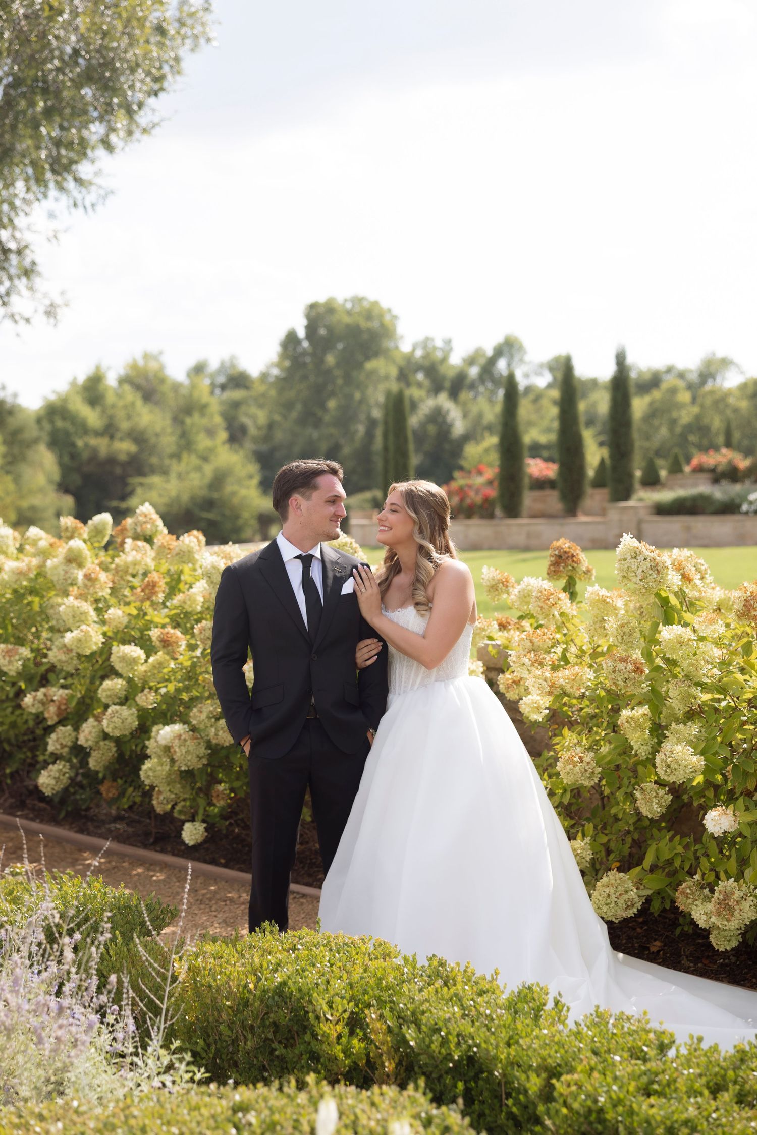 A couple in wedding attire shares a romantic moment in a garden setting with blooming hydrangea bushes.