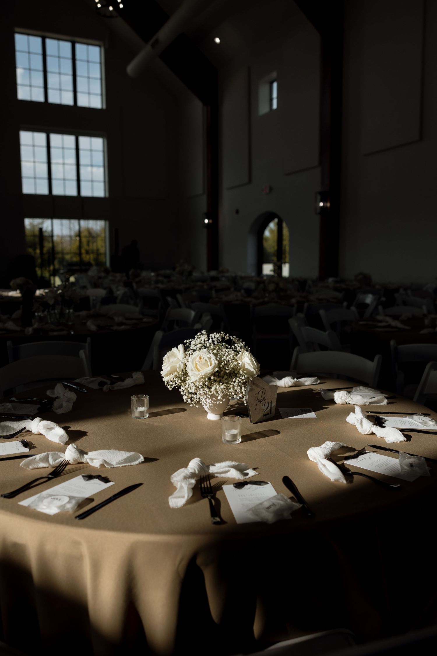 Elegant reception table setting with white floral centerpiece in a dramatic venue with high windows.