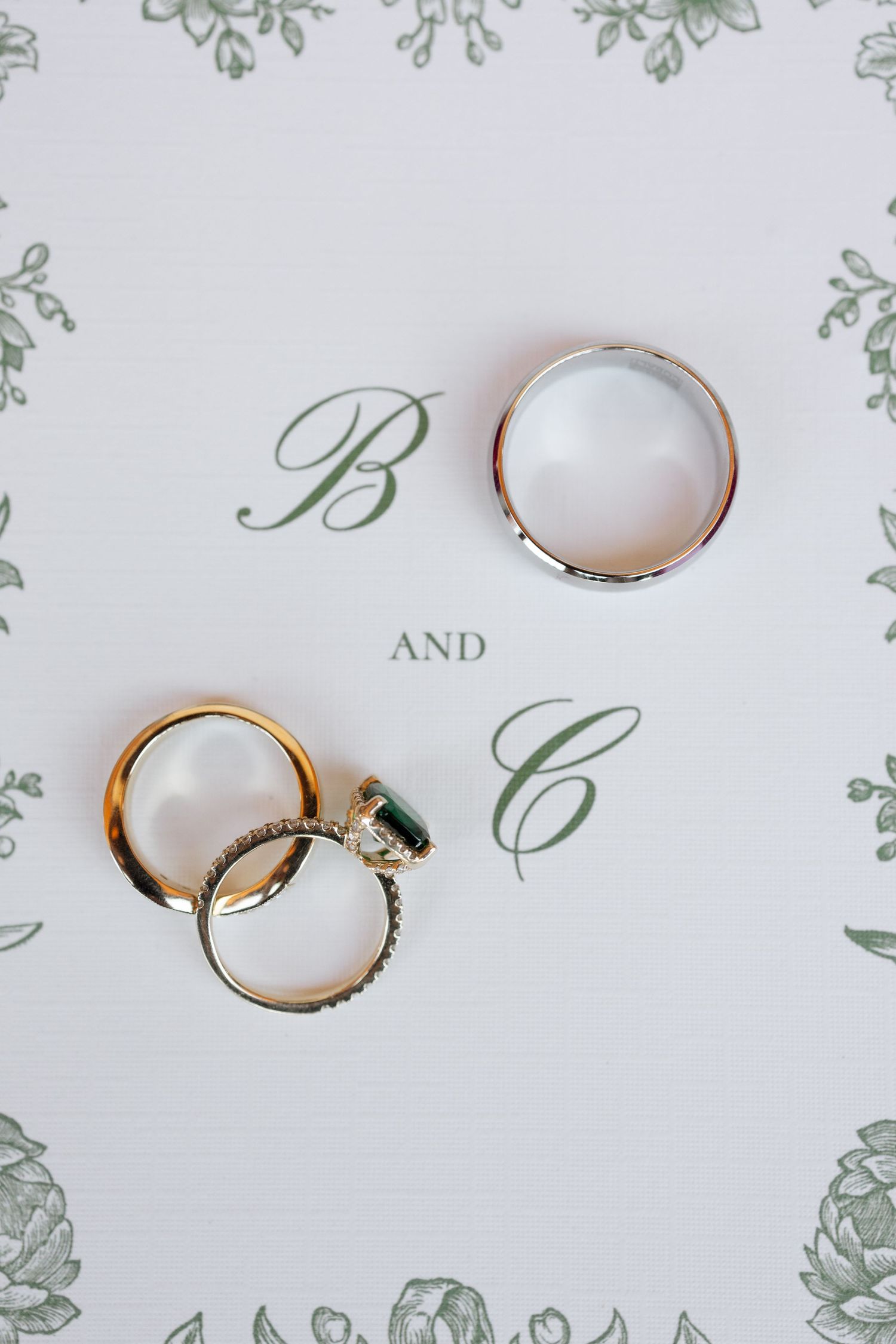 Three wedding rings arranged on white paper with decorative green monogram border.
