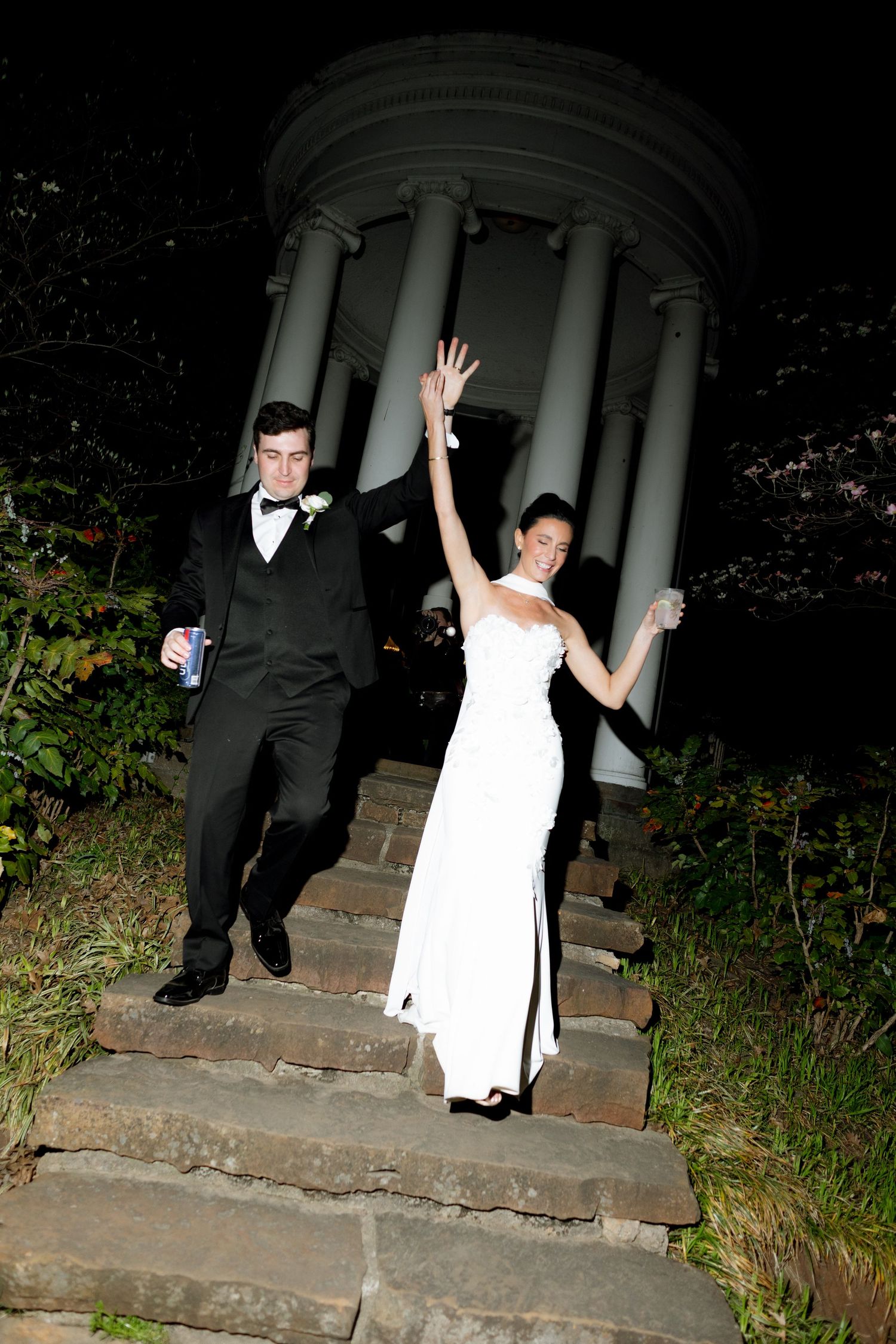 Bride in white gown raises hand joyfully as couple descends stone steps from a columned gazebo at night.