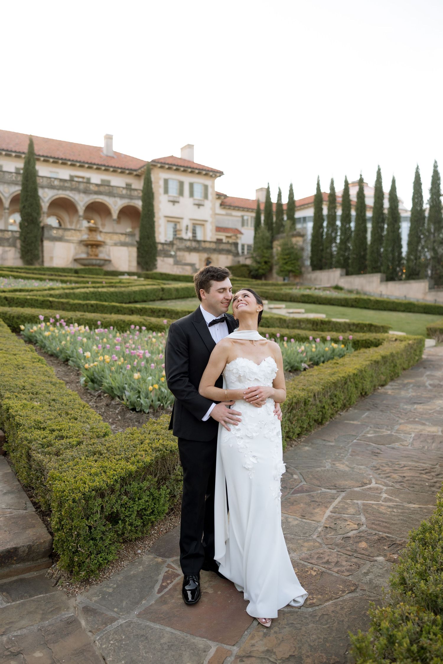 Couple embraces in formal garden with manicured hedges and tulips outside grand estate.