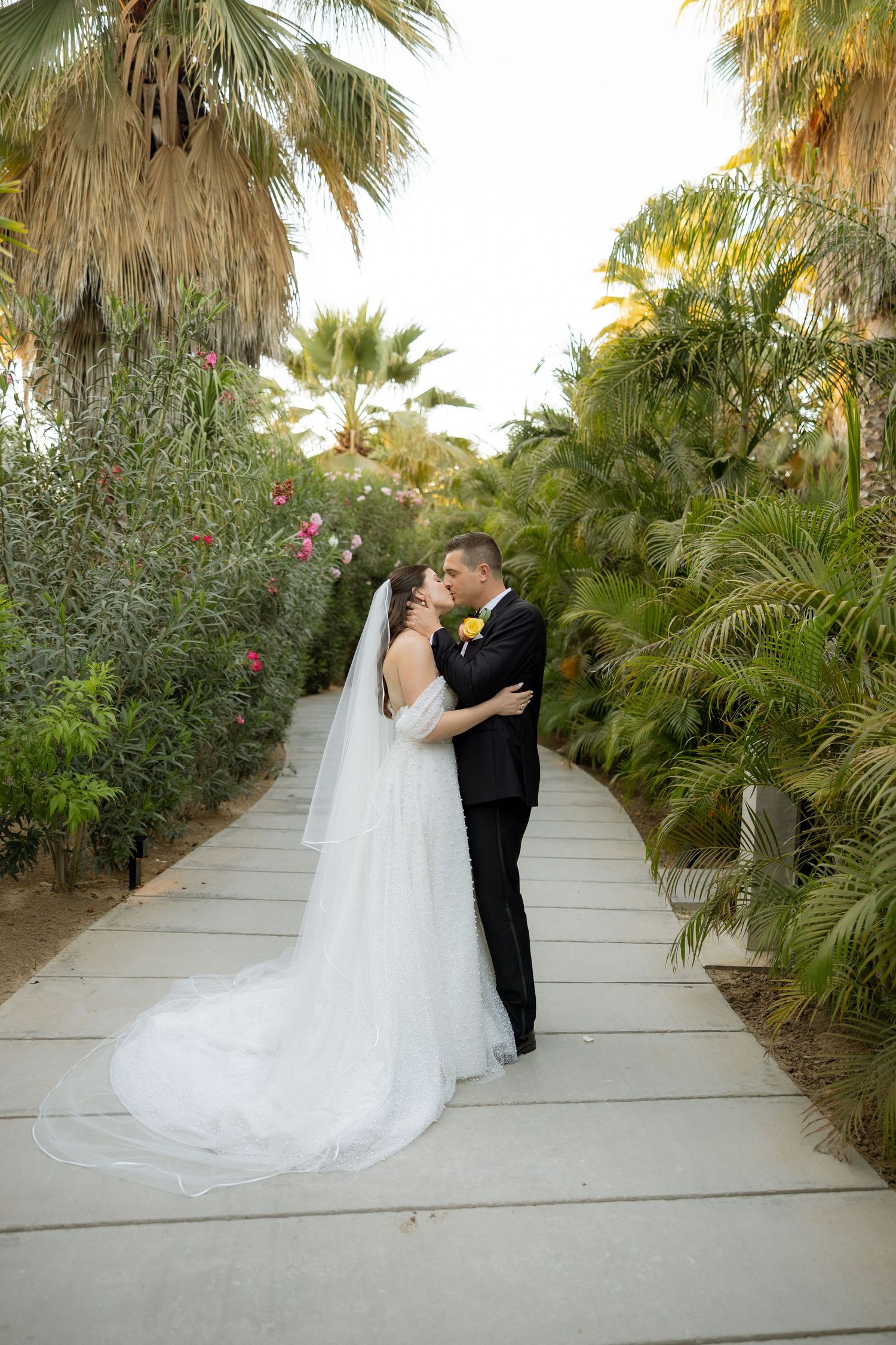 Bride and groom sharing a kiss on a garden pathway surrounded by tropical palm trees and pink flowers.