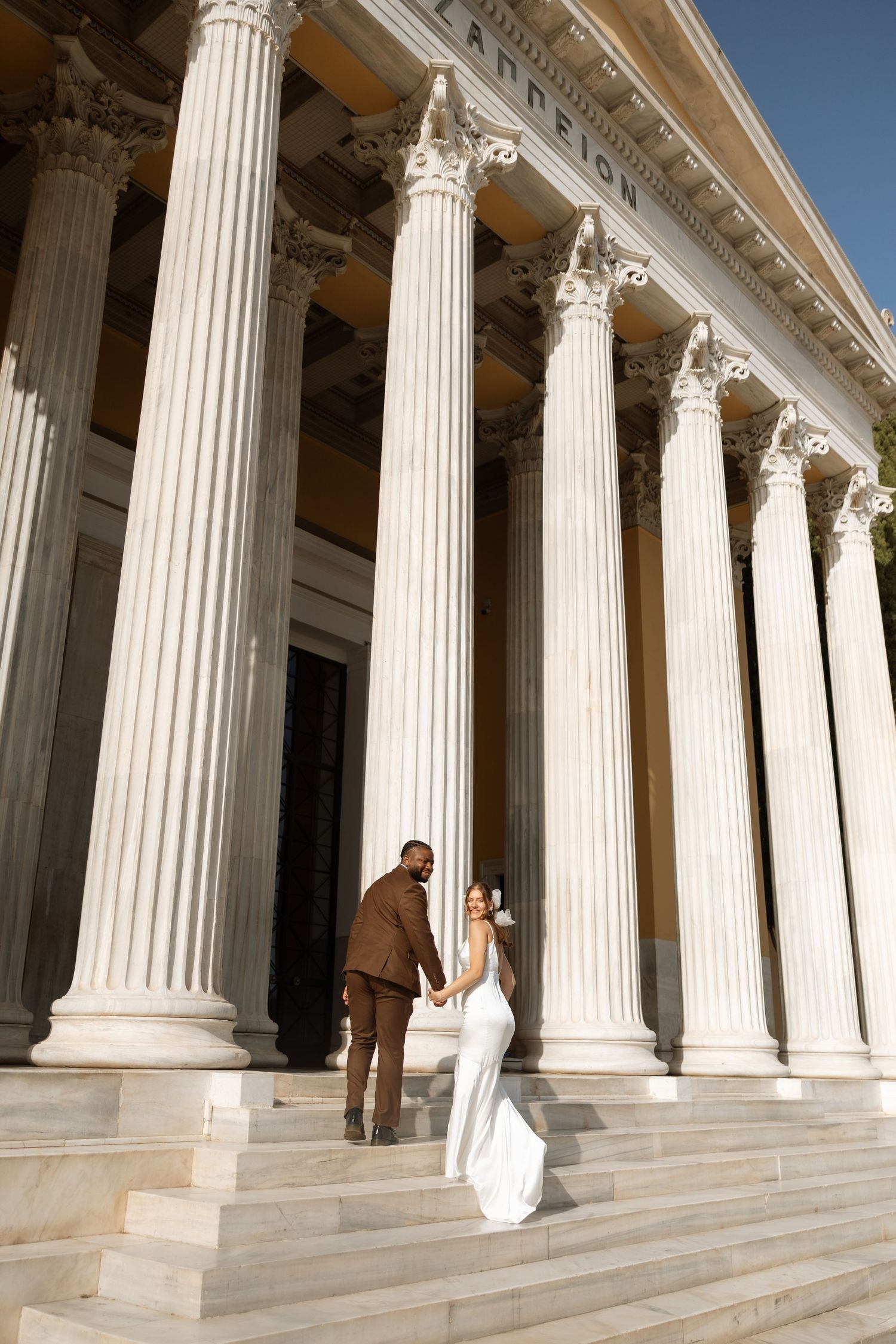 Wedding couple climbs marble steps of neoclassical building with massive columns.