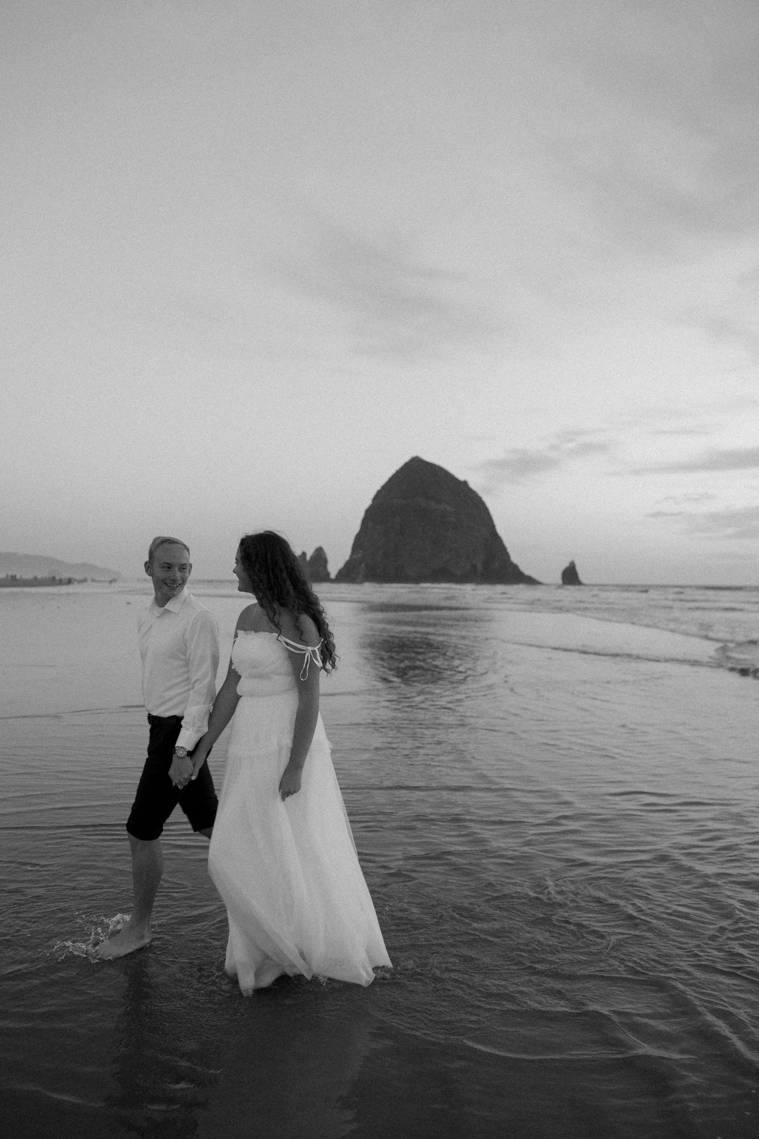 A black and white beach wedding photo with Haystack Rock silhouetted against cloudy skies.