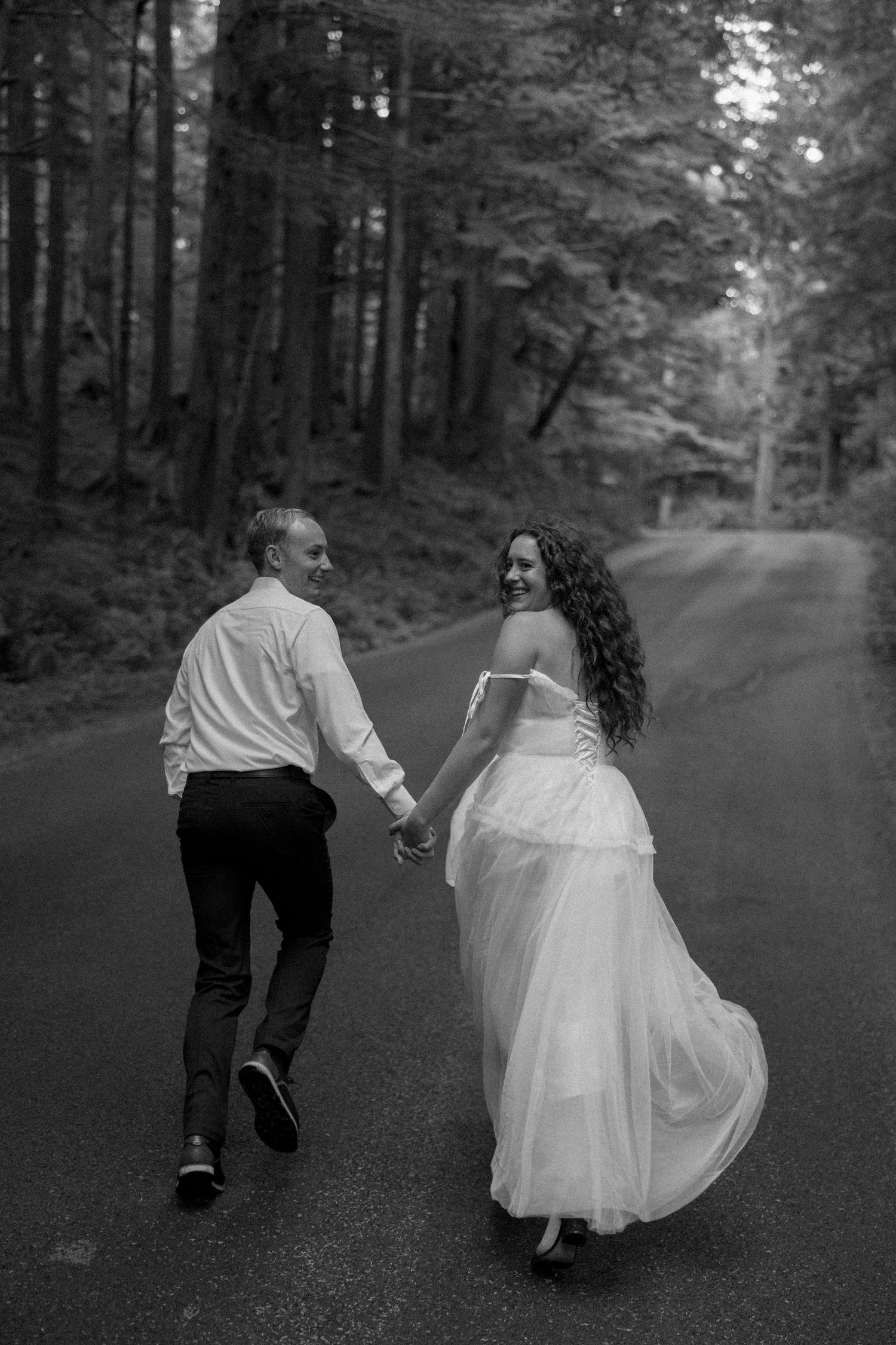 A couple holds hands while running down a forested road in black and white.