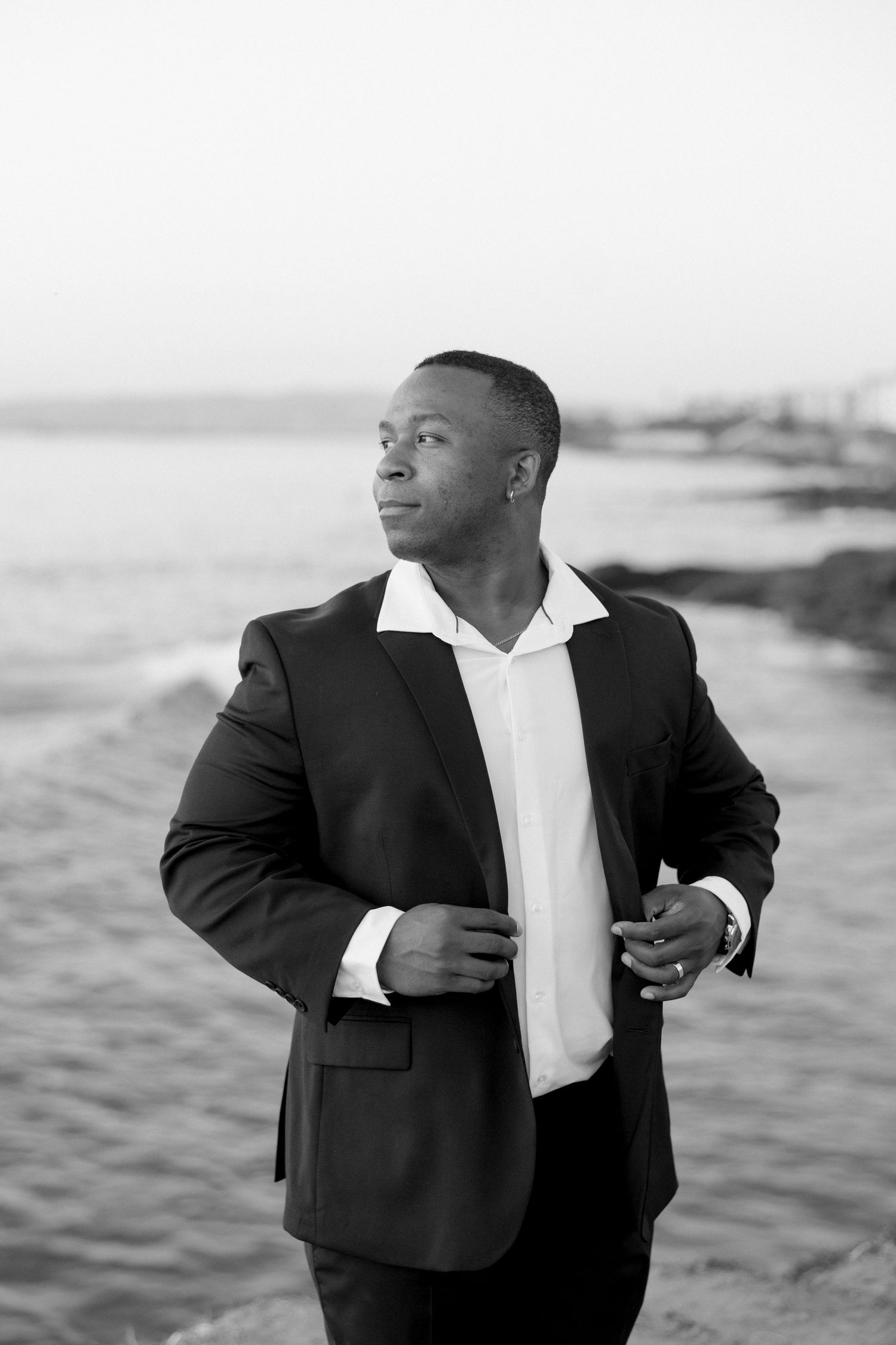 A person in a black suit and white shirt poses thoughtfully against a beach backdrop in a black and white photograph.