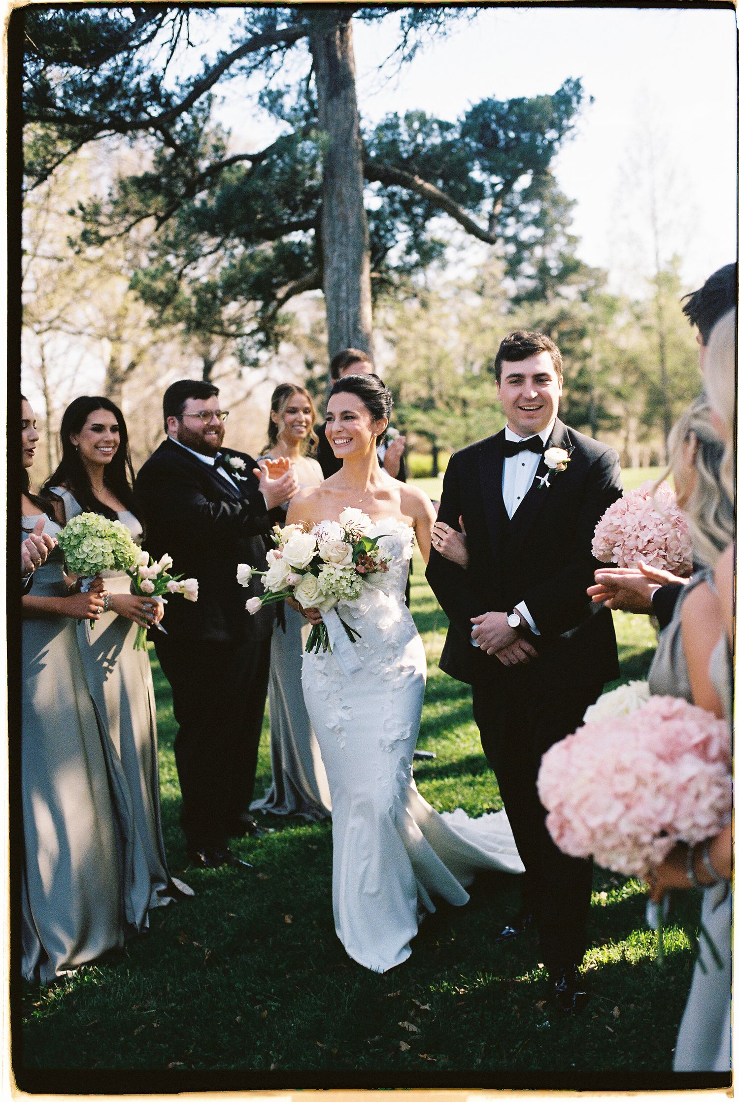 Bride and groom walk down the aisle with wedding party in sage green gowns holding pink bouquets outdoors.