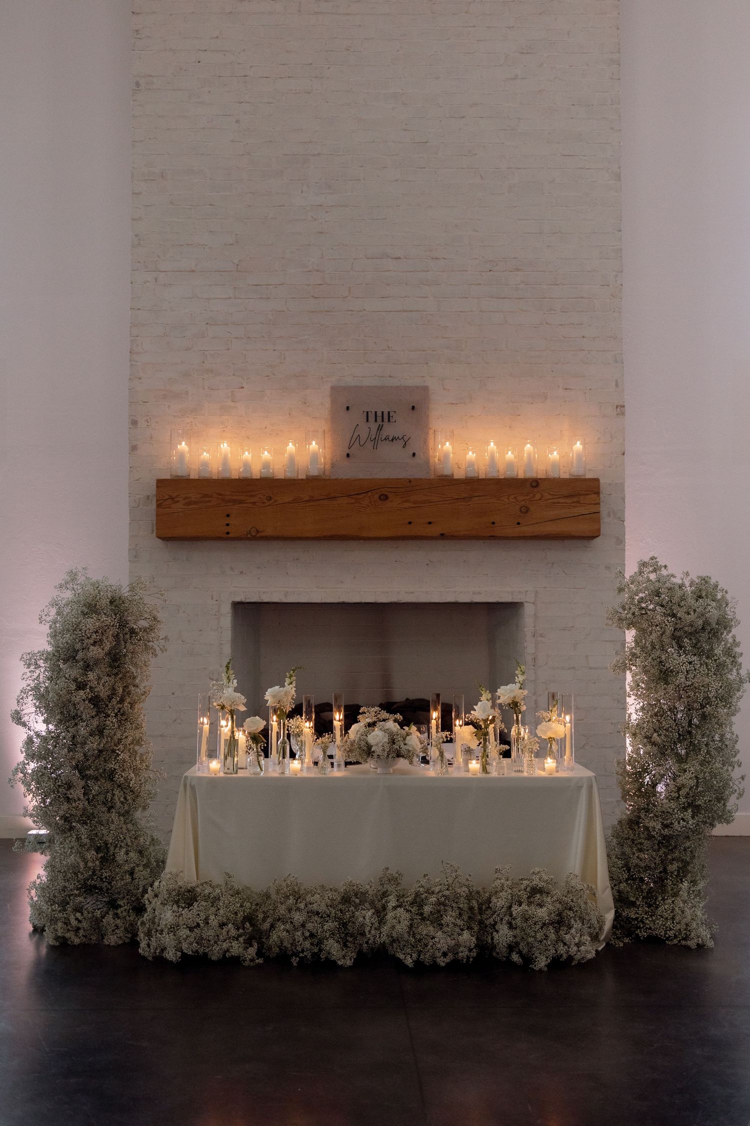 A romantic wedding reception setup with candlelit table and white floral decorations against a brick fireplace.