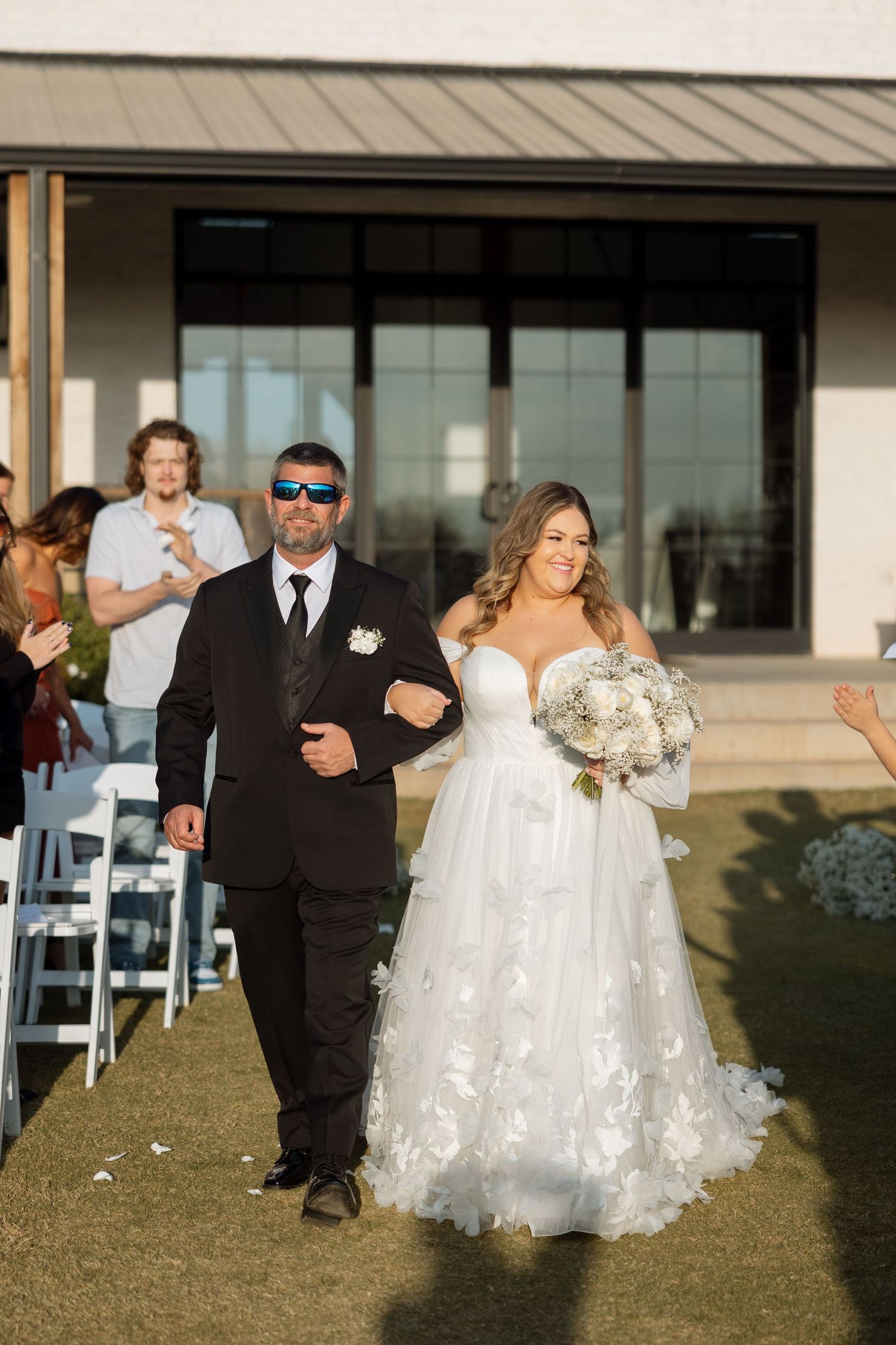 Wedding processional walking down outdoor aisle with modern building in background.