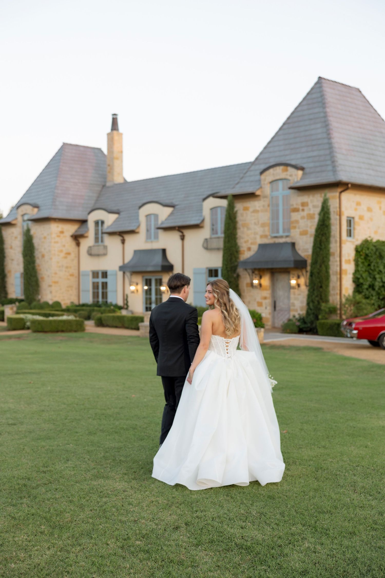 A bride and groom walk hand in hand across a green lawn with a stone estate building in the background.