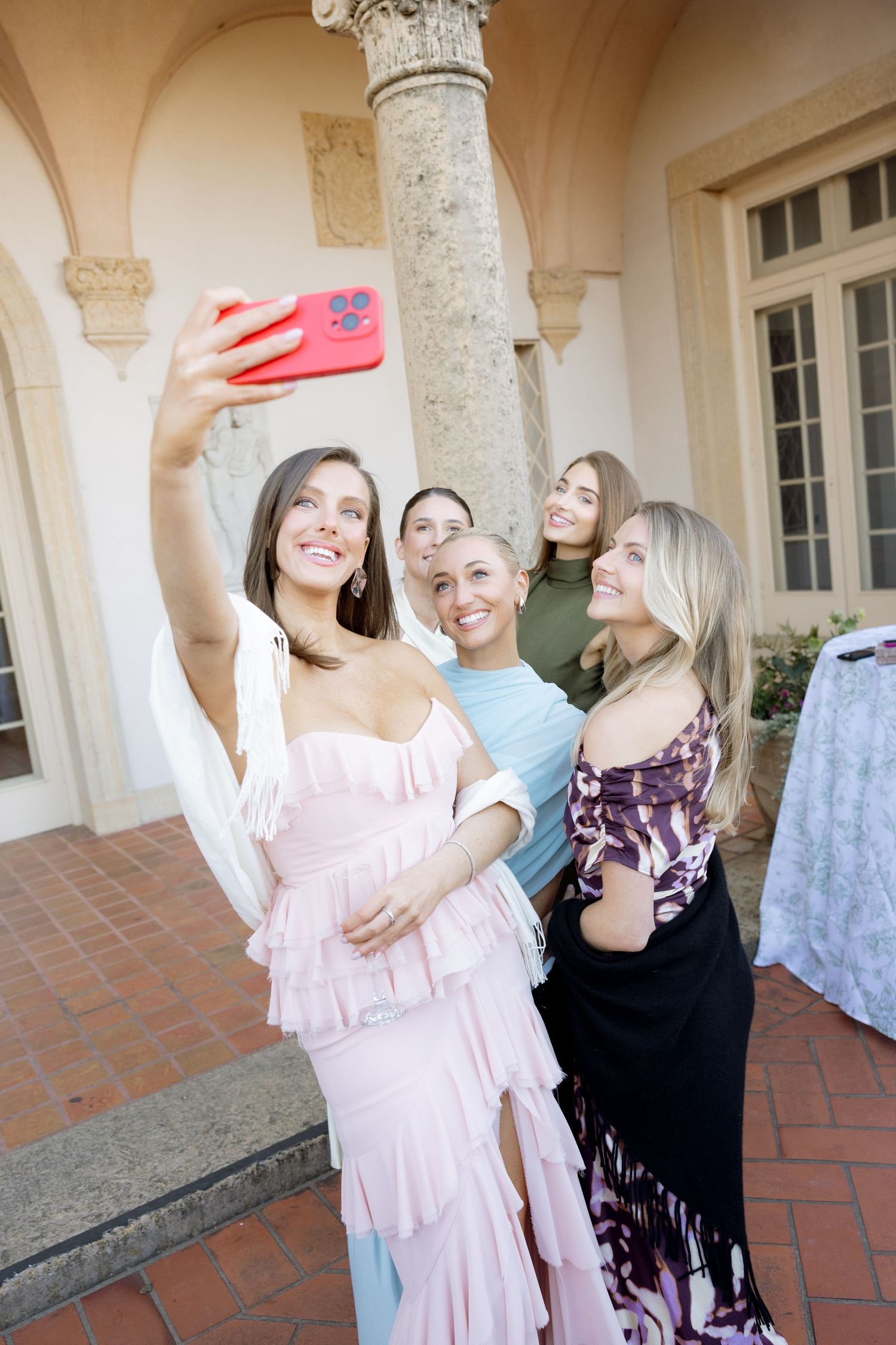Four women in formal dresses taking a selfie with red phone at wedding venue.