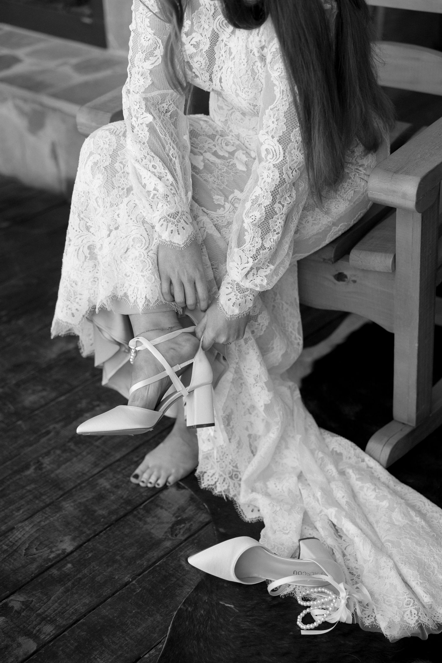Close-up of bride putting on white wedding shoes, wearing lace dress.