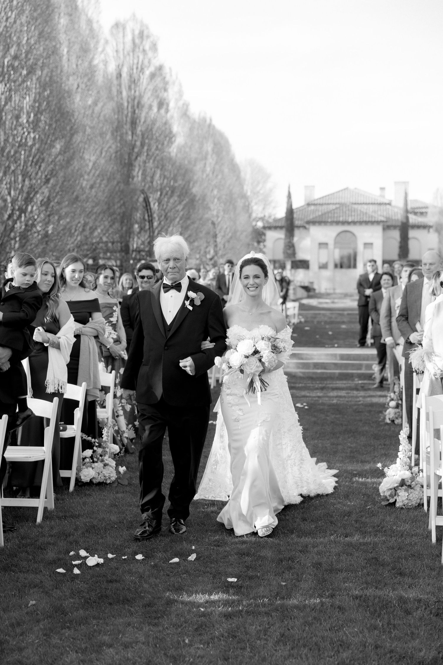 Black and white photo of bride and father walking down flower-lined aisle at outdoor wedding.