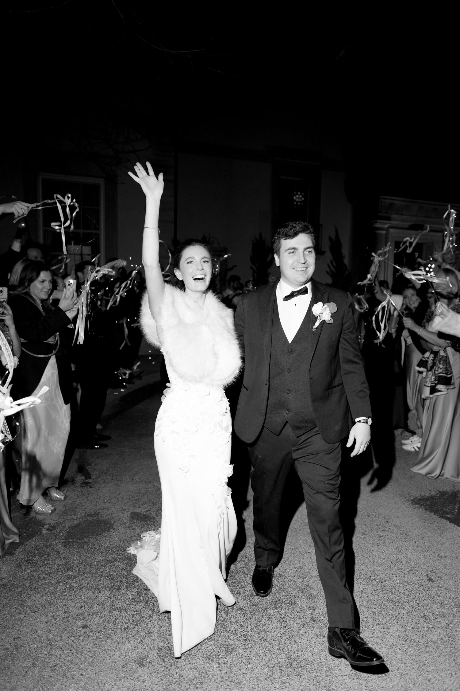 Joyful bride and groom exit wedding reception in black and white photo, bride waving to cheering guests.