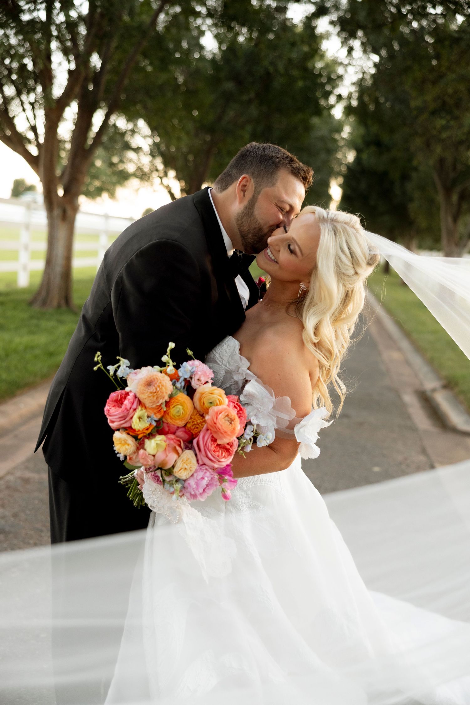 Romantic wedding couple embracing on a tree-lined path with a vibrant floral bouquet.