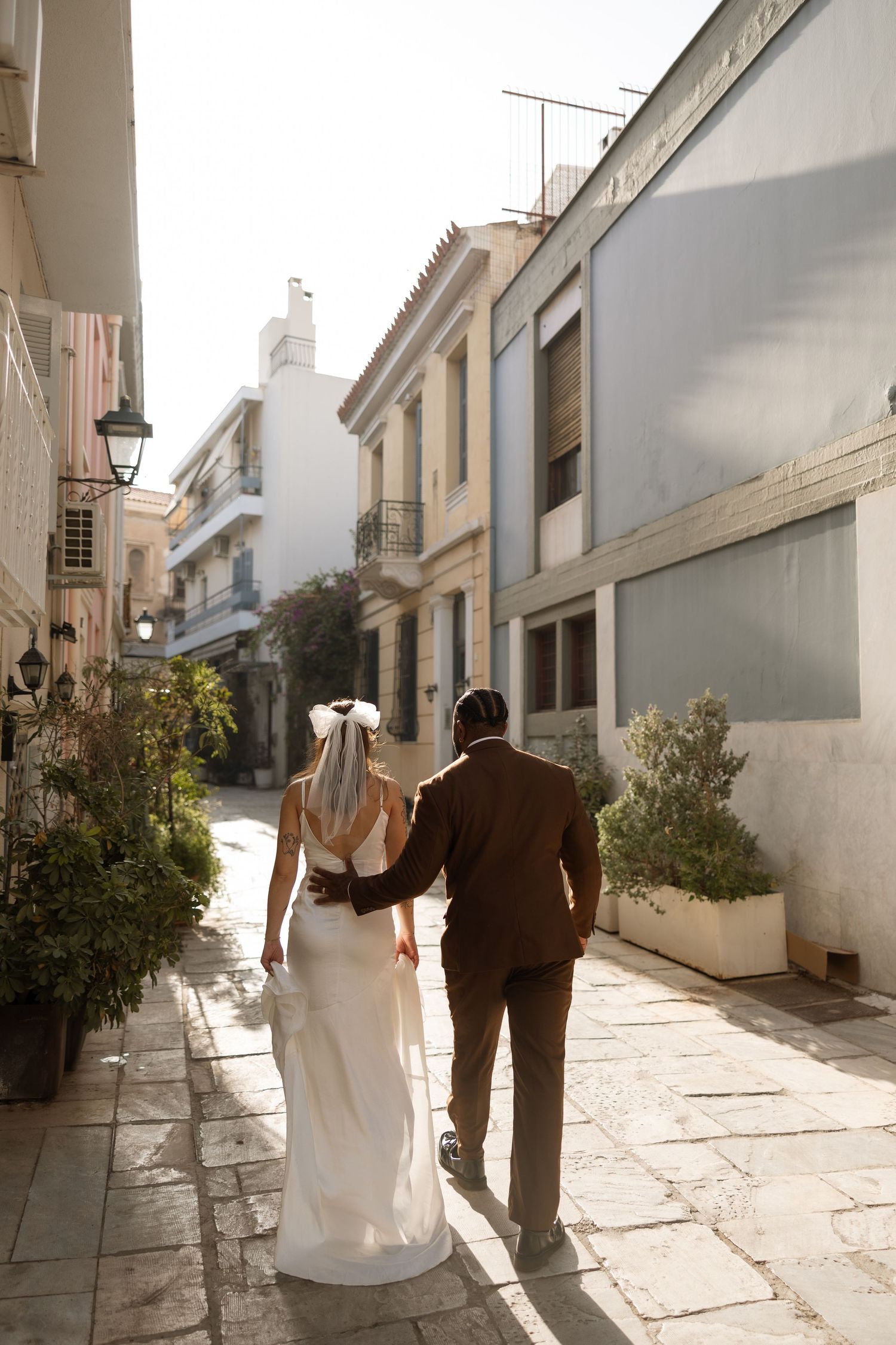 Couple walking hand in hand down a sunlit Mediterranean alley with white buildings.