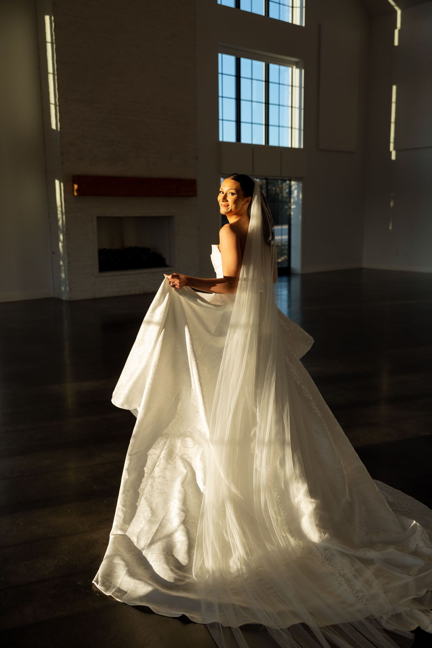 A flowing white wedding gown with long veil is illuminated dramatically against tall windows in an elegant venue.