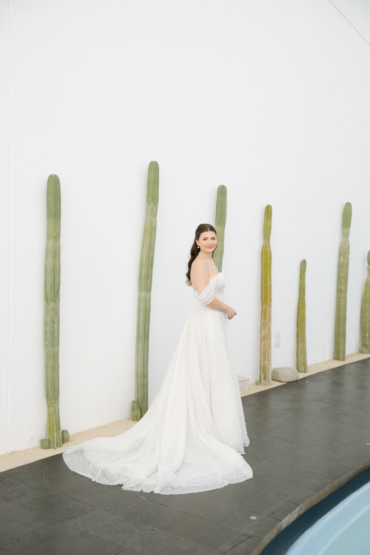 Elegant bride in off-shoulder white gown with train poses by white wall with tall cacti beside a pool.