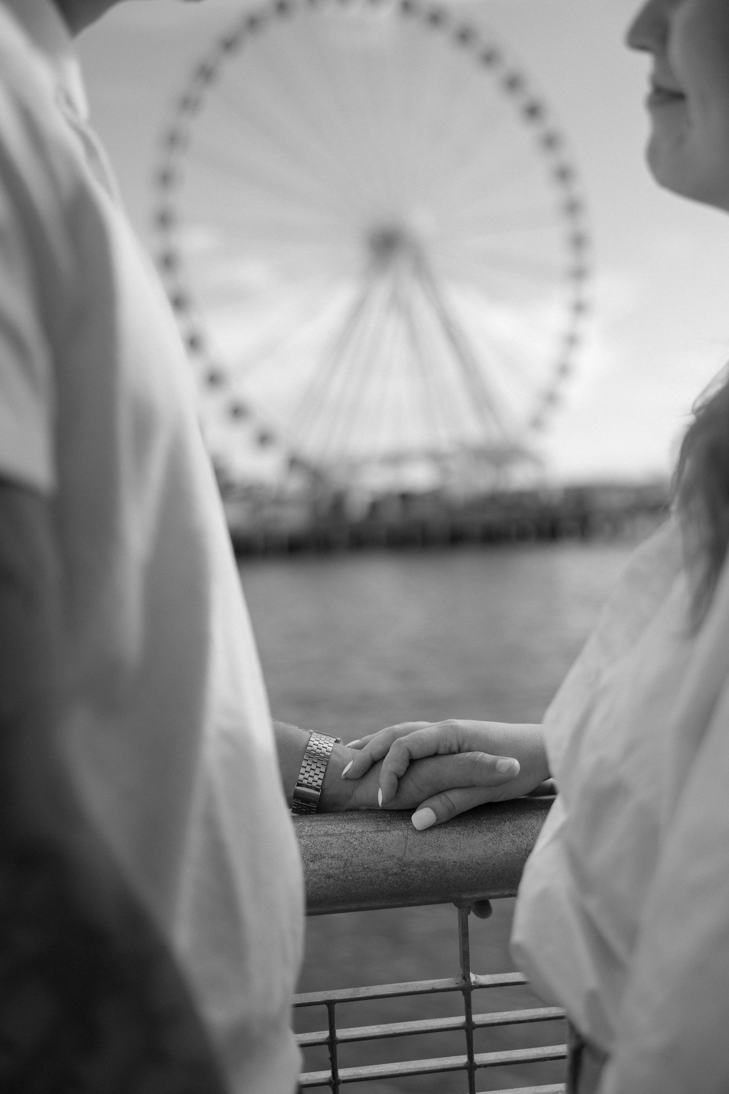 Black and white detail shot of hands resting on railing with Ferris wheel in background.