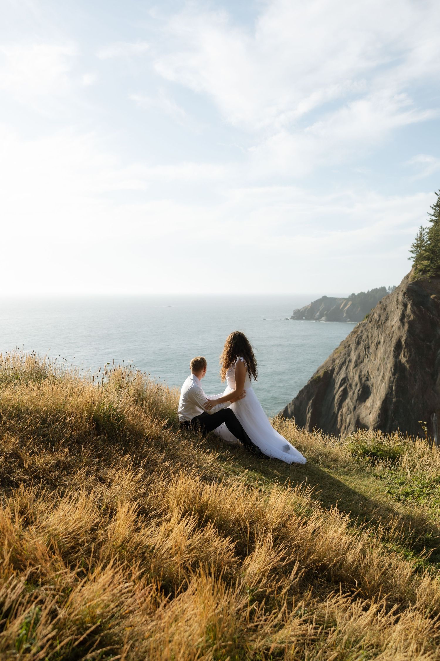 Wedding couple sits on golden grass overlooking dramatic coastal cliffs and ocean waves.