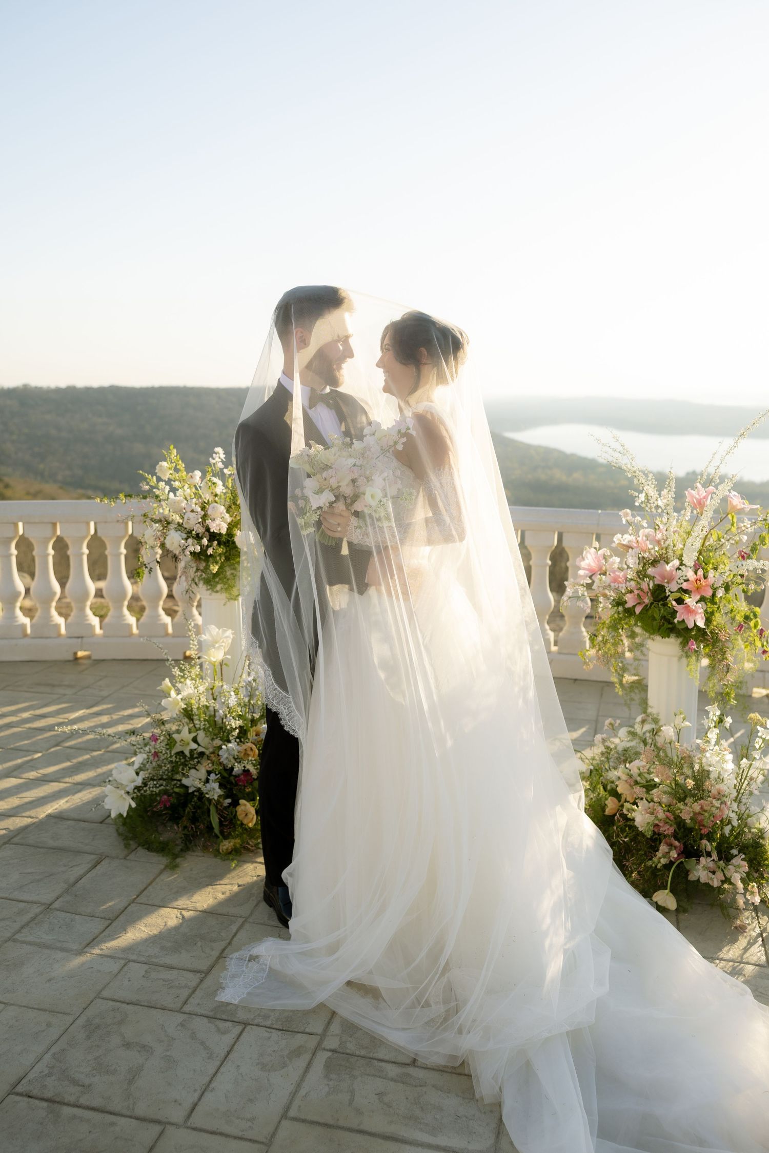 Bride and groom share a romantic moment under a veil on a terrace with floral arrangements and scenic views.