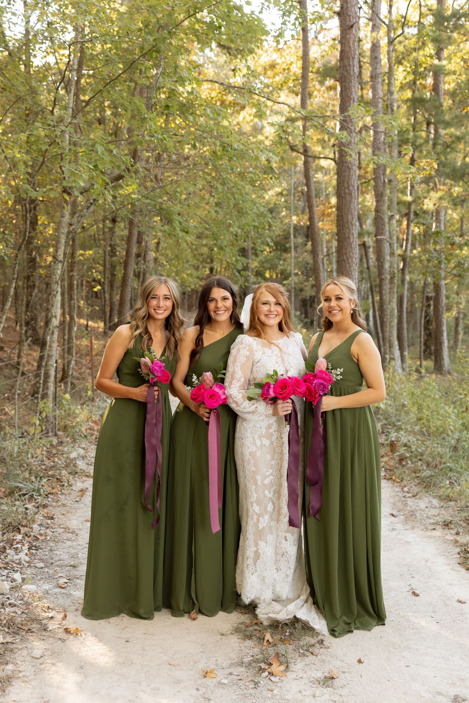 Wedding party with bridesmaids in olive green dresses holding bright pink bouquets on forest path.