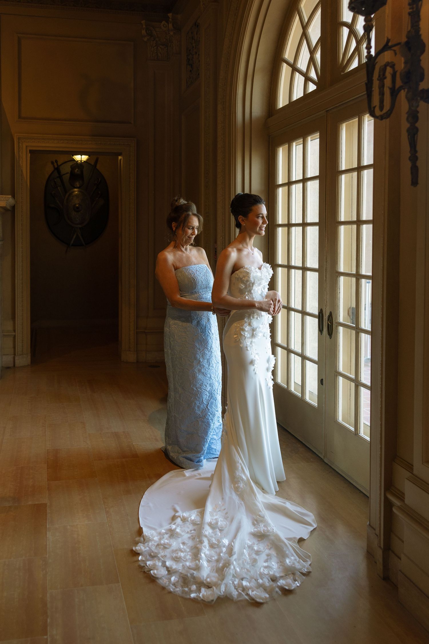 Bride in white gown with floral train stands with bridesmaid in blue dress by elegant French doors.