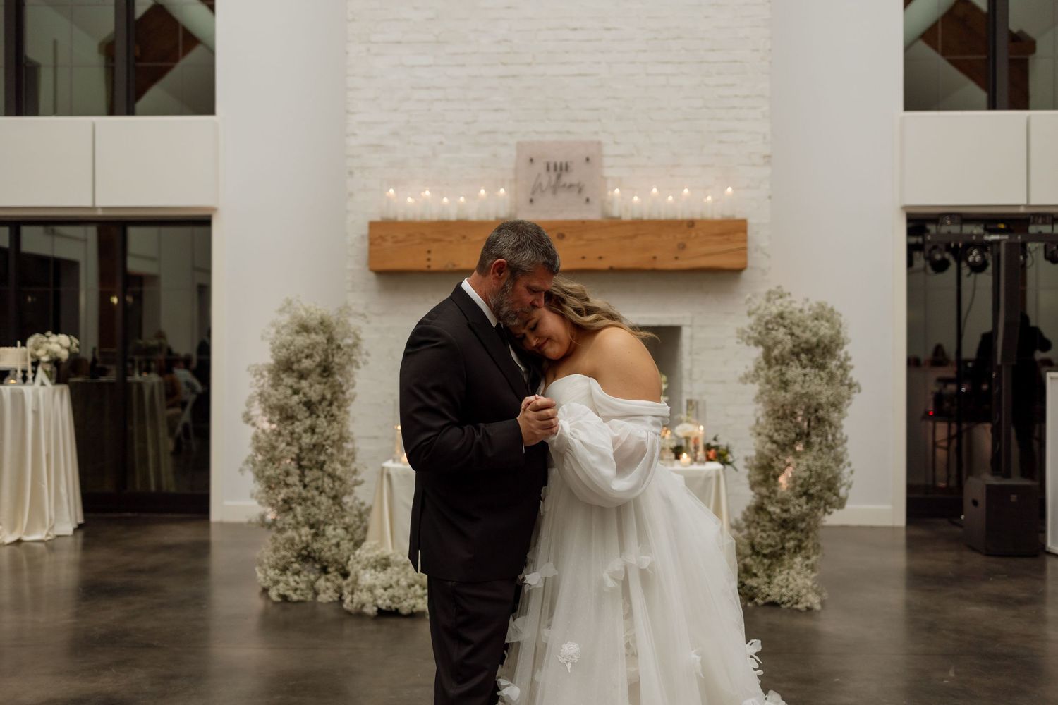 A couple shares an intimate moment at their wedding ceremony in front of a white brick fireplace with floral decorations.