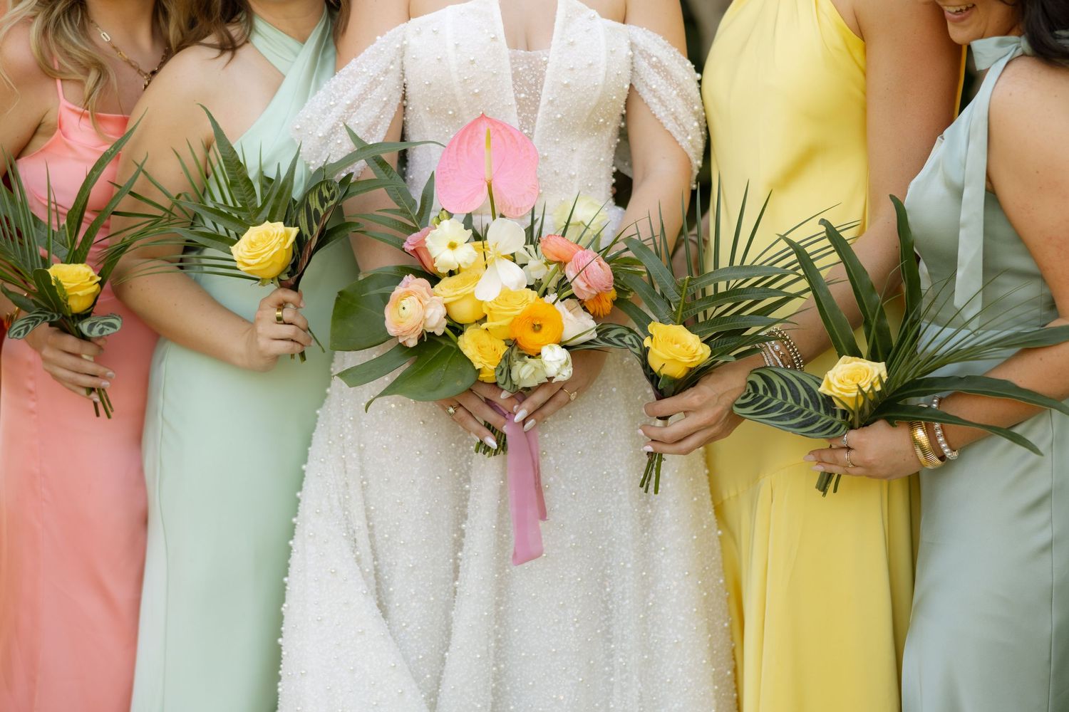 Bride and bridesmaids in pastel dresses holding tropical bouquets with yellow roses and palm leaves.