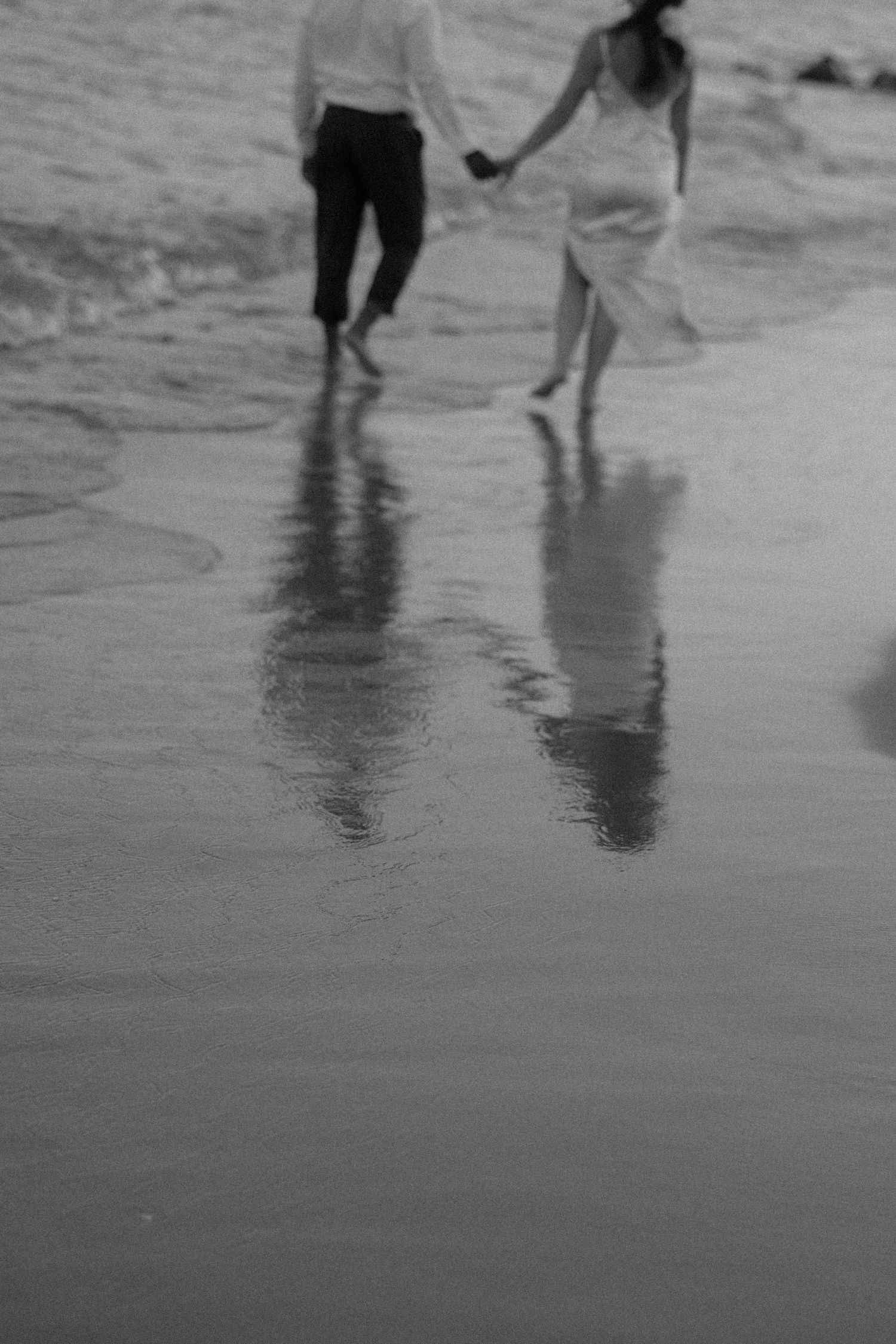 Black and white image of feet walking along a wet beach shore with reflections in the sand.