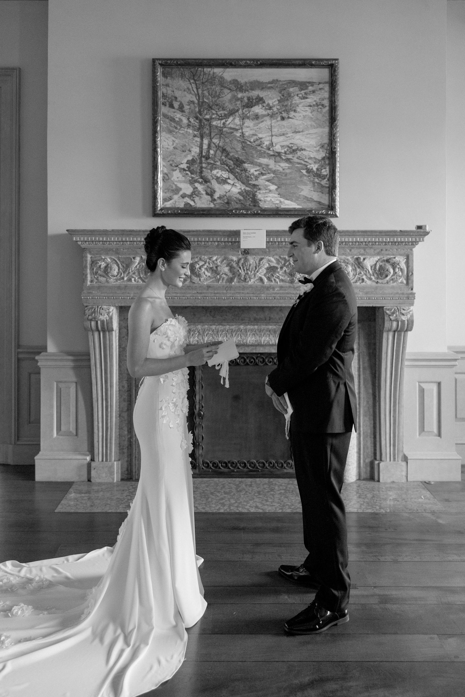 Black and white first look photo of bride and groom standing by ornate fireplace in elegant room.