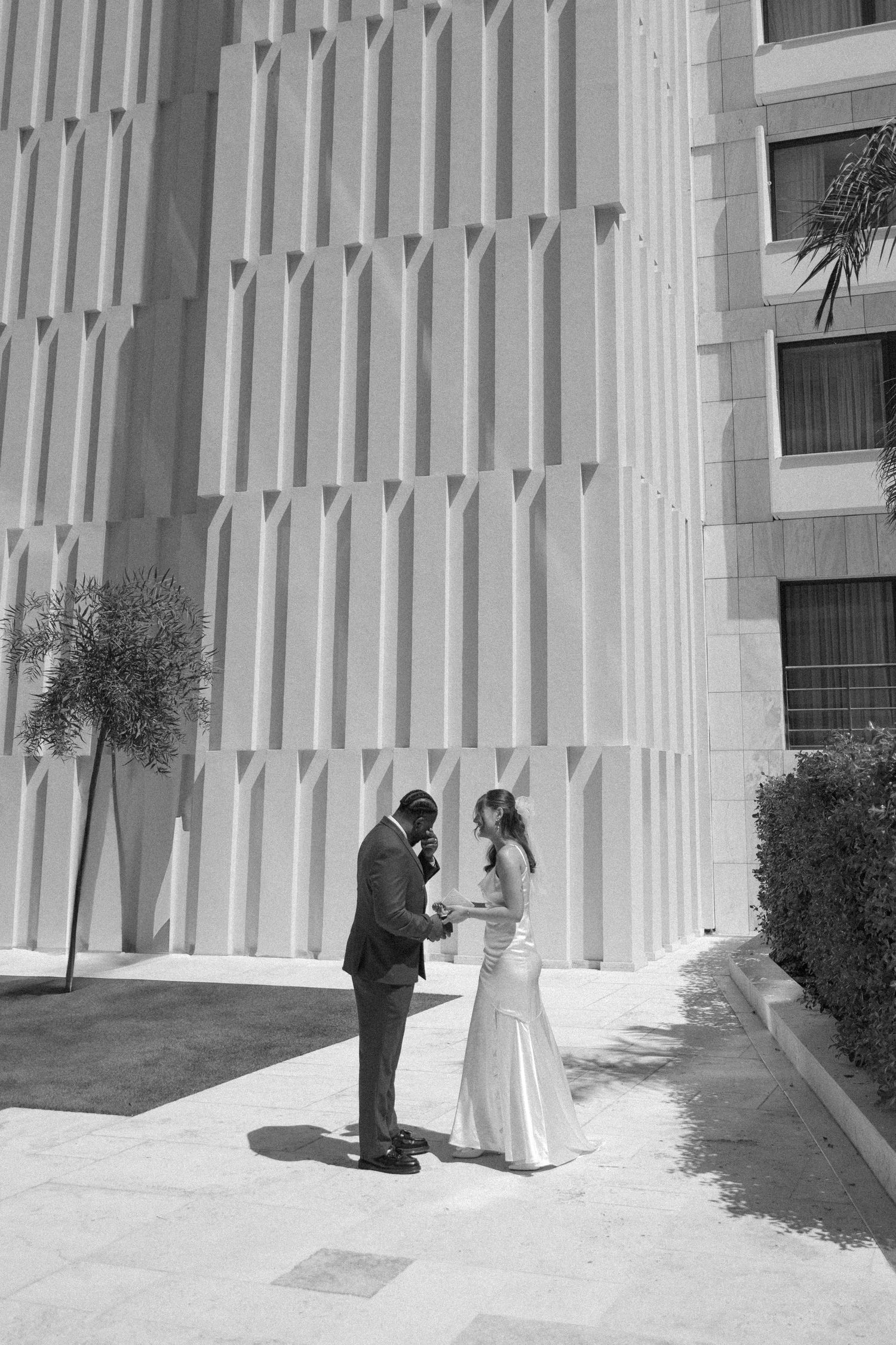 A black and white photo of a couple reading their vows beside a geometric white wall.