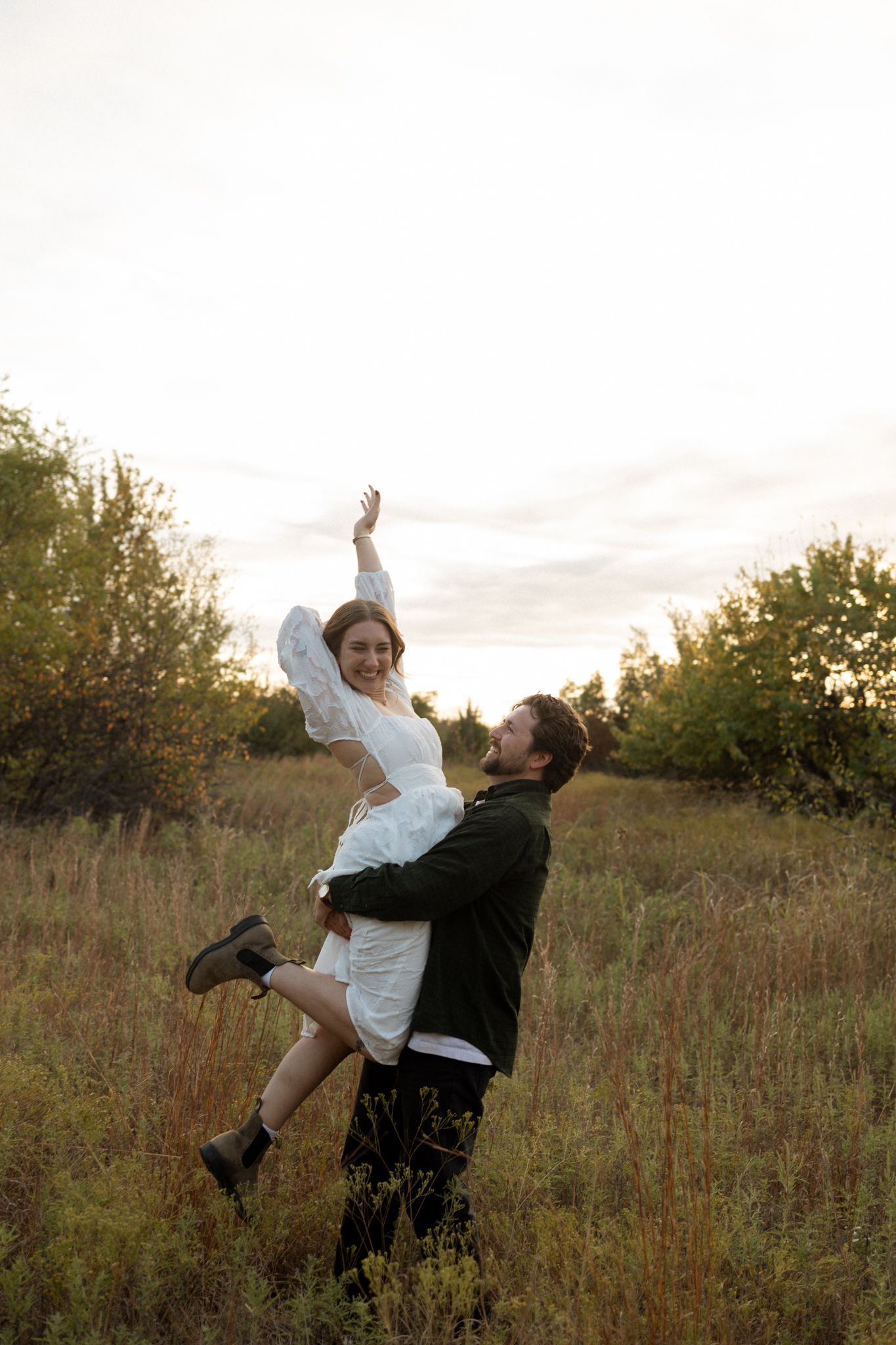 A couple in a field at sunset share a joyful moment as one person lifts the other wearing a white dress.