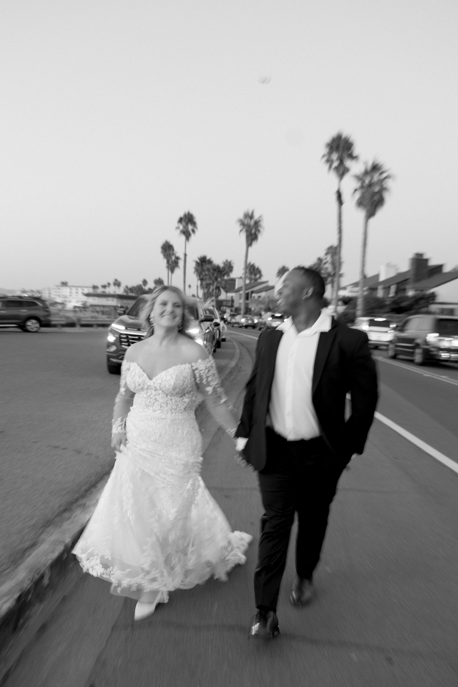 A couple in wedding attire walks along a palm tree-lined street in a series of black and white photographs.