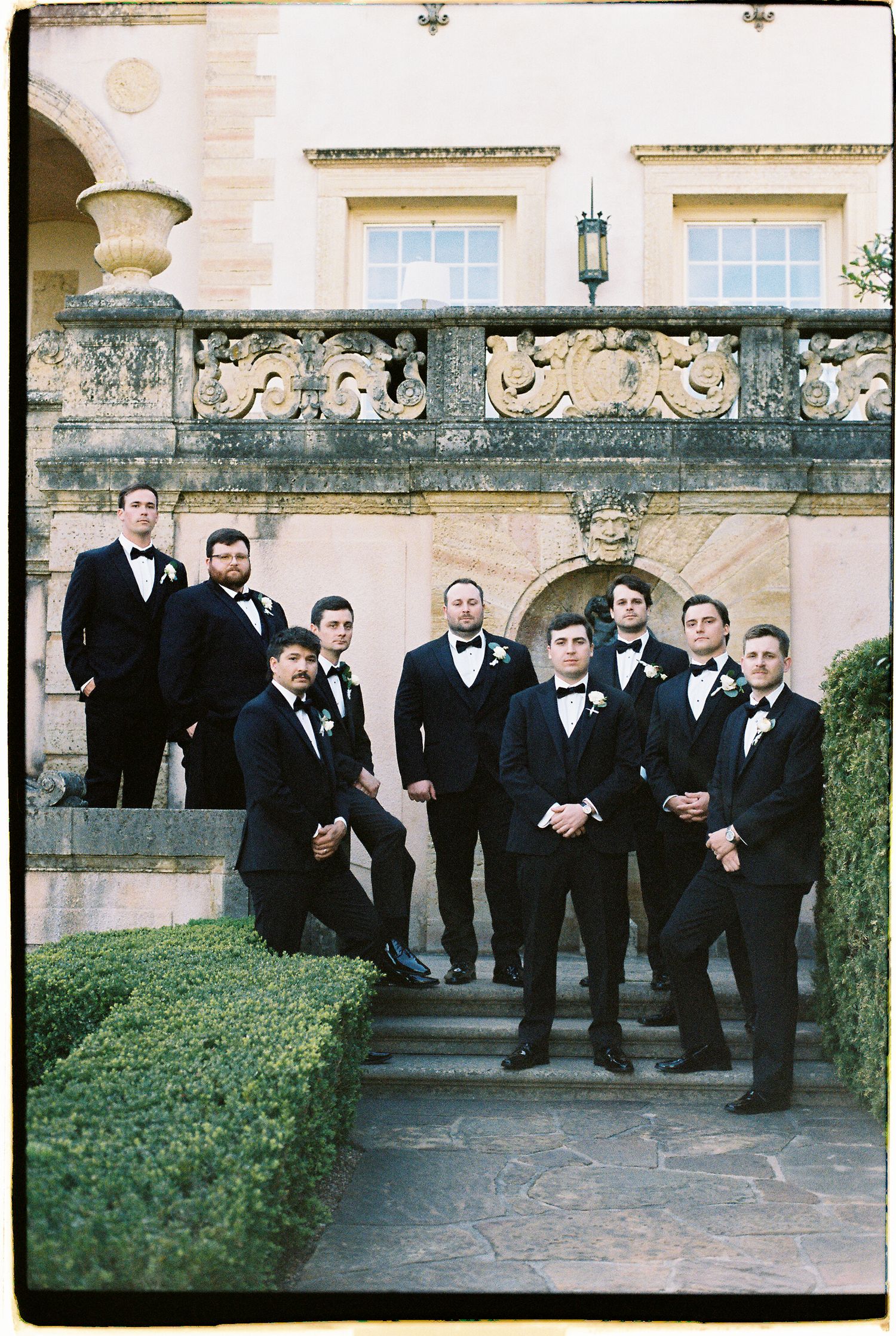 Groom and groomsmen in classic black tuxedos posing on stone steps of elegant estate venue.
