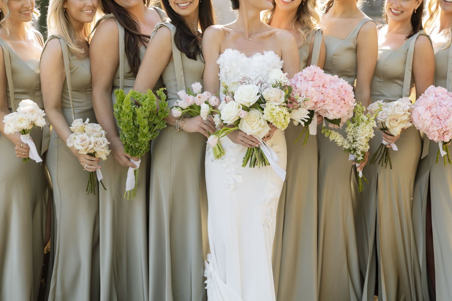 Bride in white lace gown with bridesmaids in sage green dresses holding pink and white bouquets.