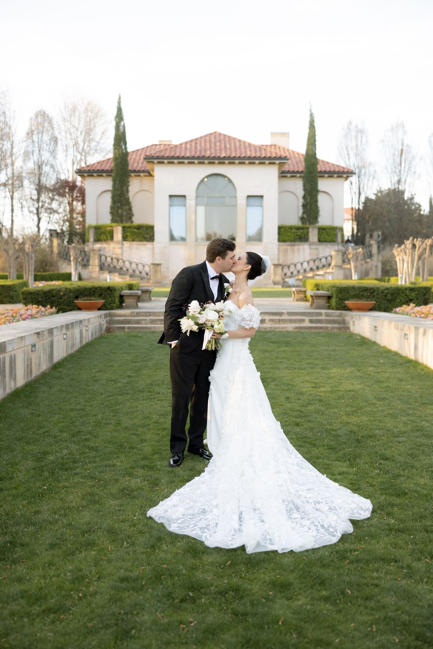 Bride and groom share a kiss on manicured lawn before Italian-style villa with cypress trees.
