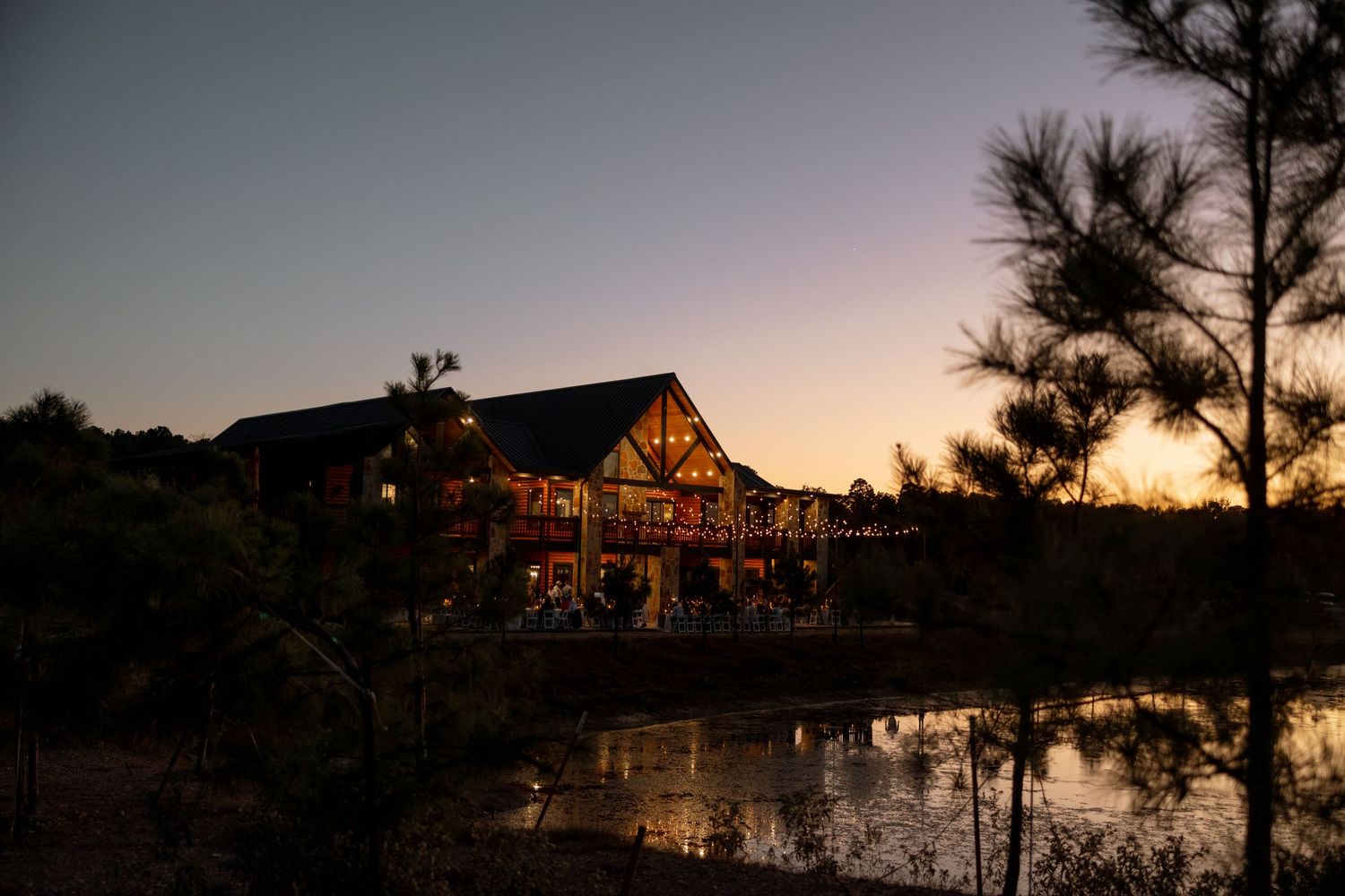 Rustic log cabin lodge overlooking a tranquil pond at sunset with pine trees silhouetted against the twilight sky.